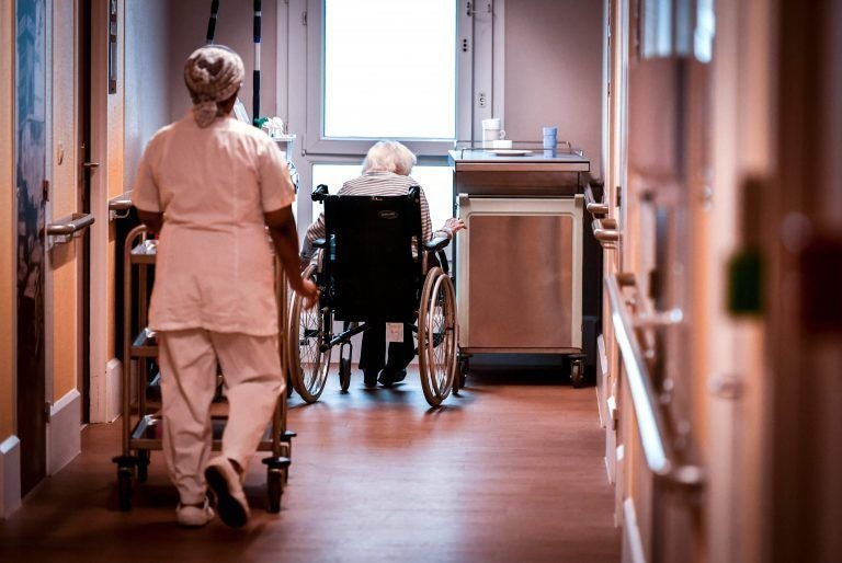 Eine Pflegerin schiebt einen Rollwagen auf einen Flur mit einer Seniorin im Rollstuhl.