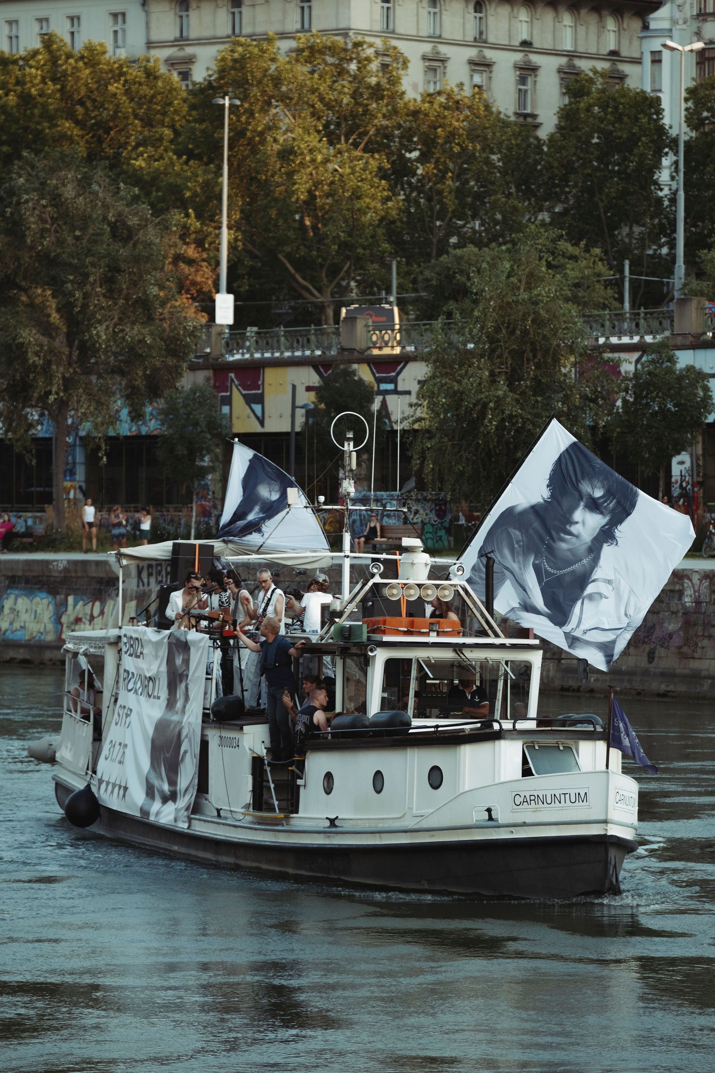 Ein Boot mit großen Flaggen und Porträts fährt einen Fluss entlang, mehrere Menschen an Bord.