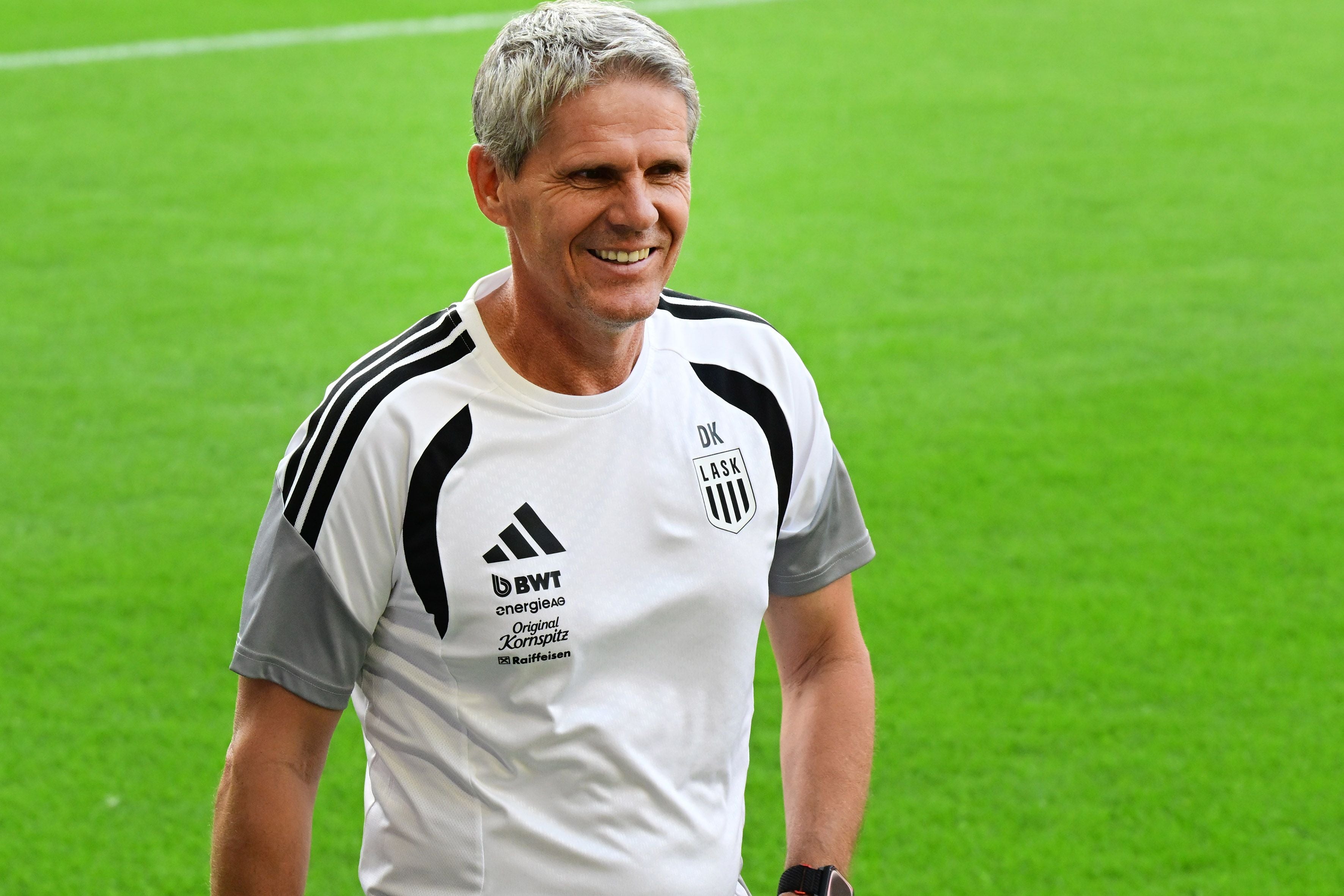 Ein lächelnder Mann im LASK-Trainertrikot steht auf einem Fußballfeld.