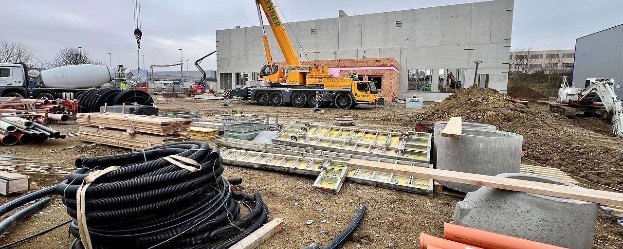 Baustelle mit Kran, Baumaterialien und Rohren vor einem halbfertigen Gebäude.
