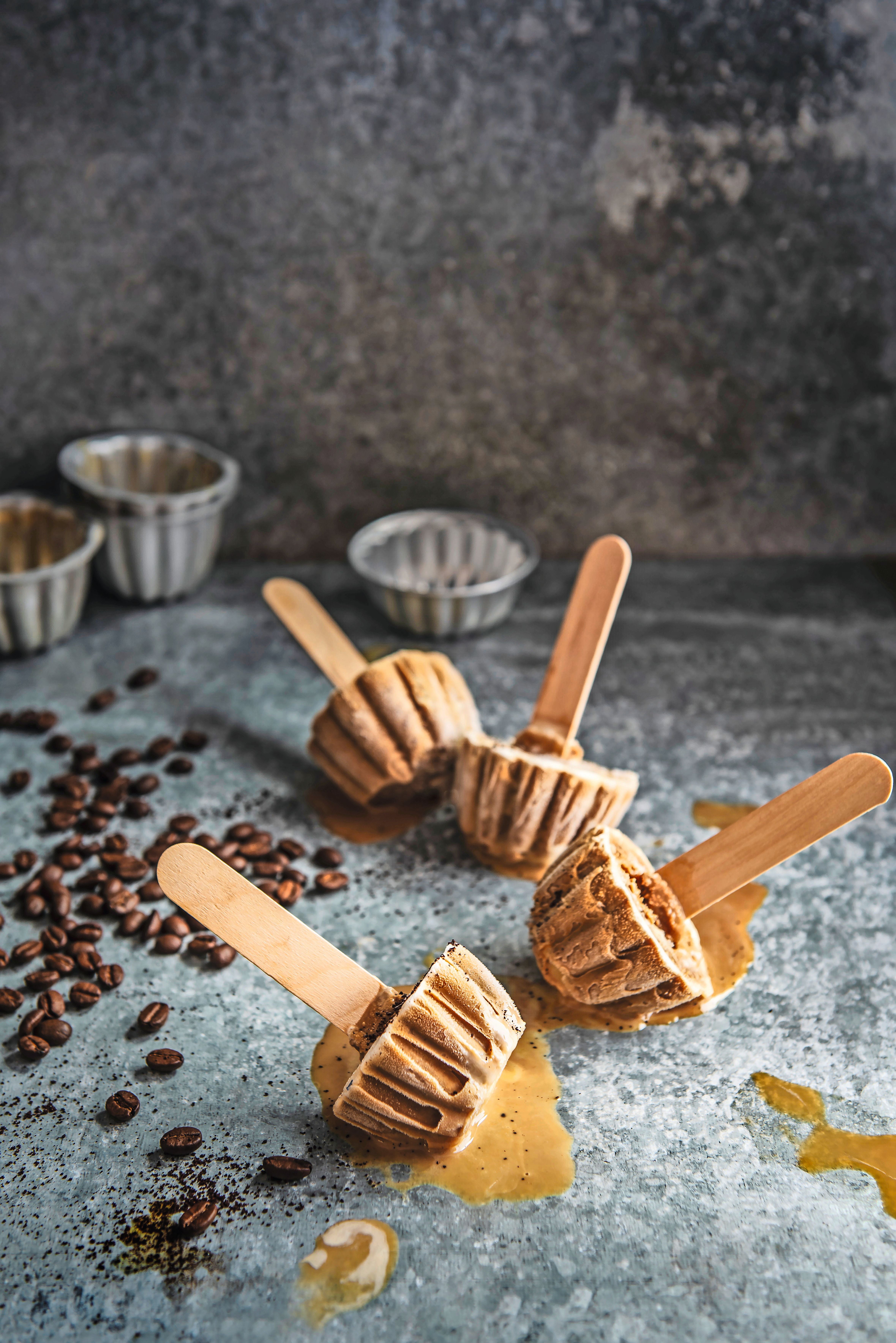 Vier Eiskaffee-Lollies in Kuchenform schmelzen auf einer Steinplatte, Kaffeebohnen liegen daneben.