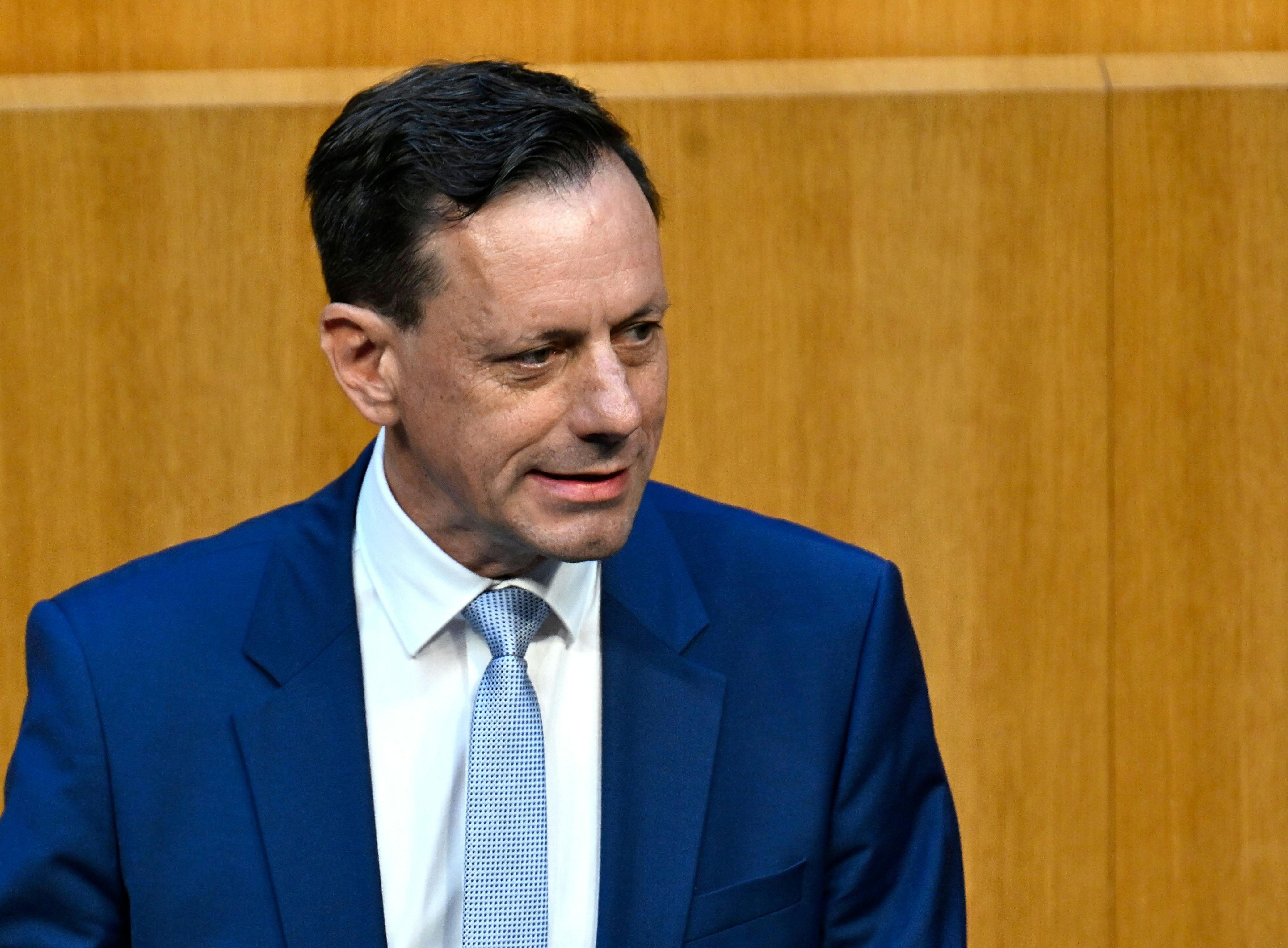 Ein Mann im blauen Anzug steht vor einer Holzvertäfelung und schaut zur Seite.