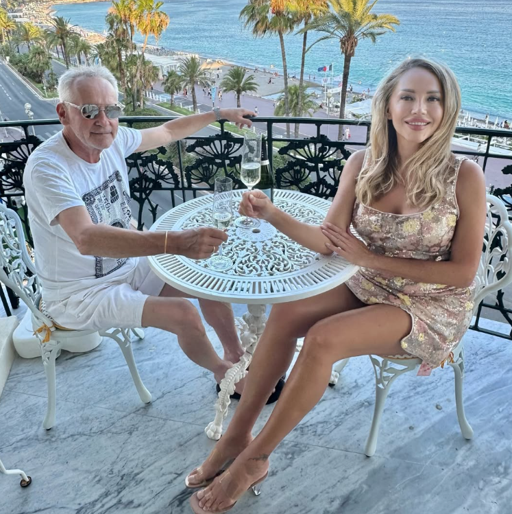 Ein älterer Mann und eine junge Frau stoßen auf einem Balkon am Meer mit Champagner an.
