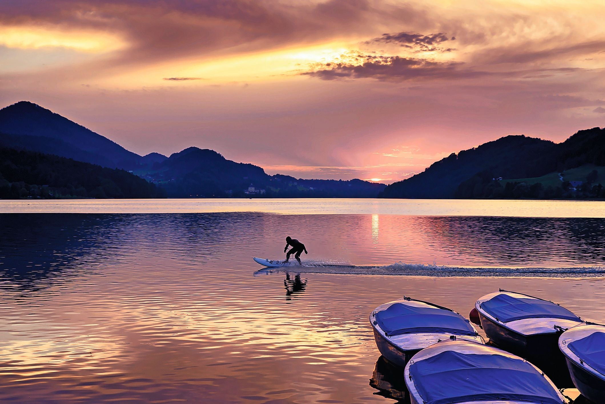 Sonnenuntergang am Fuschlsee mit Surfer auf E-Brett und Booten im Vordergrund, Berge im Hintergrund.