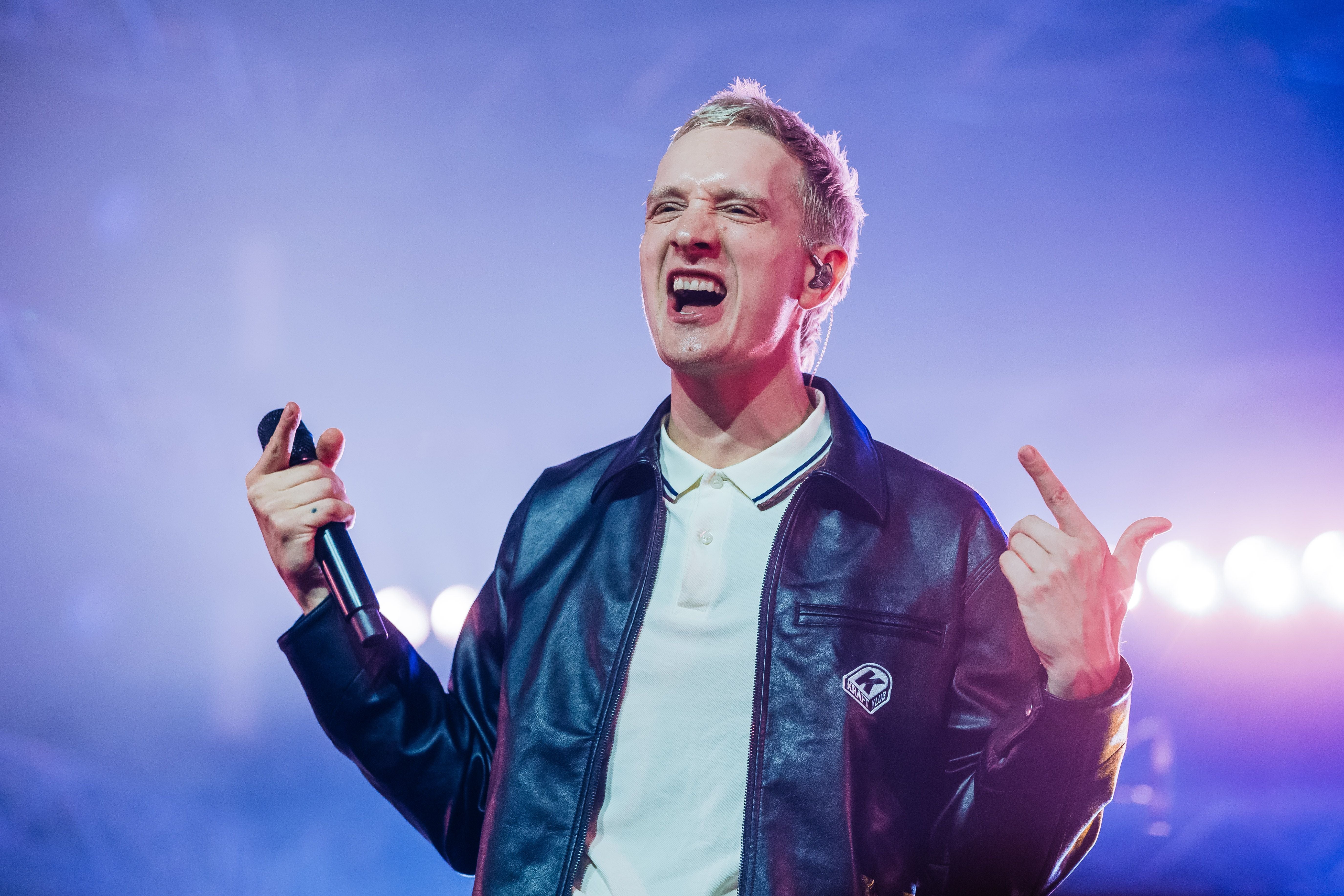 Ein Sänger mit Lederjacke und Mikrofon singt auf einer beleuchteten Bühne.