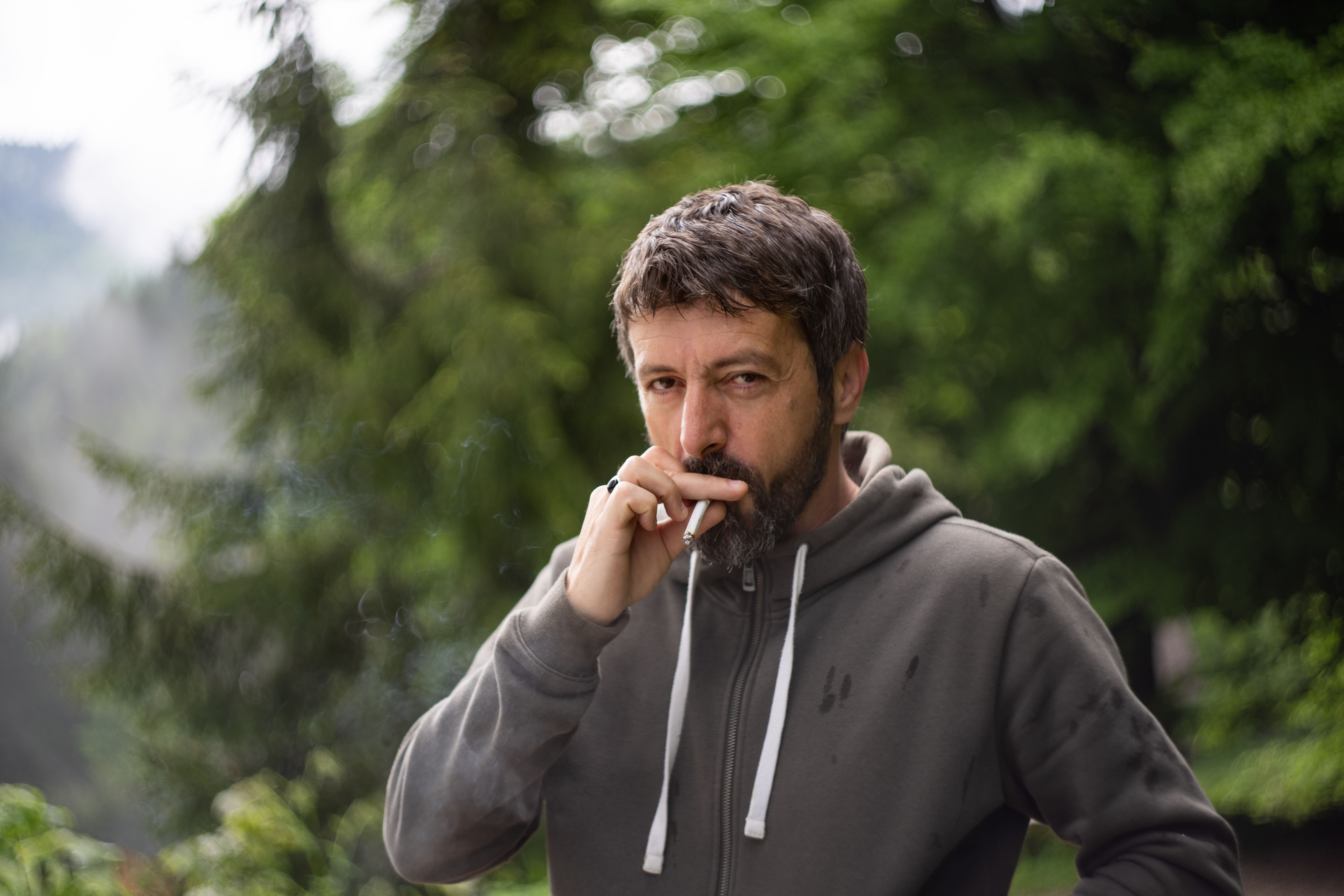 Mann mit grauem Hoodie raucht eine Zigarette im Wald.