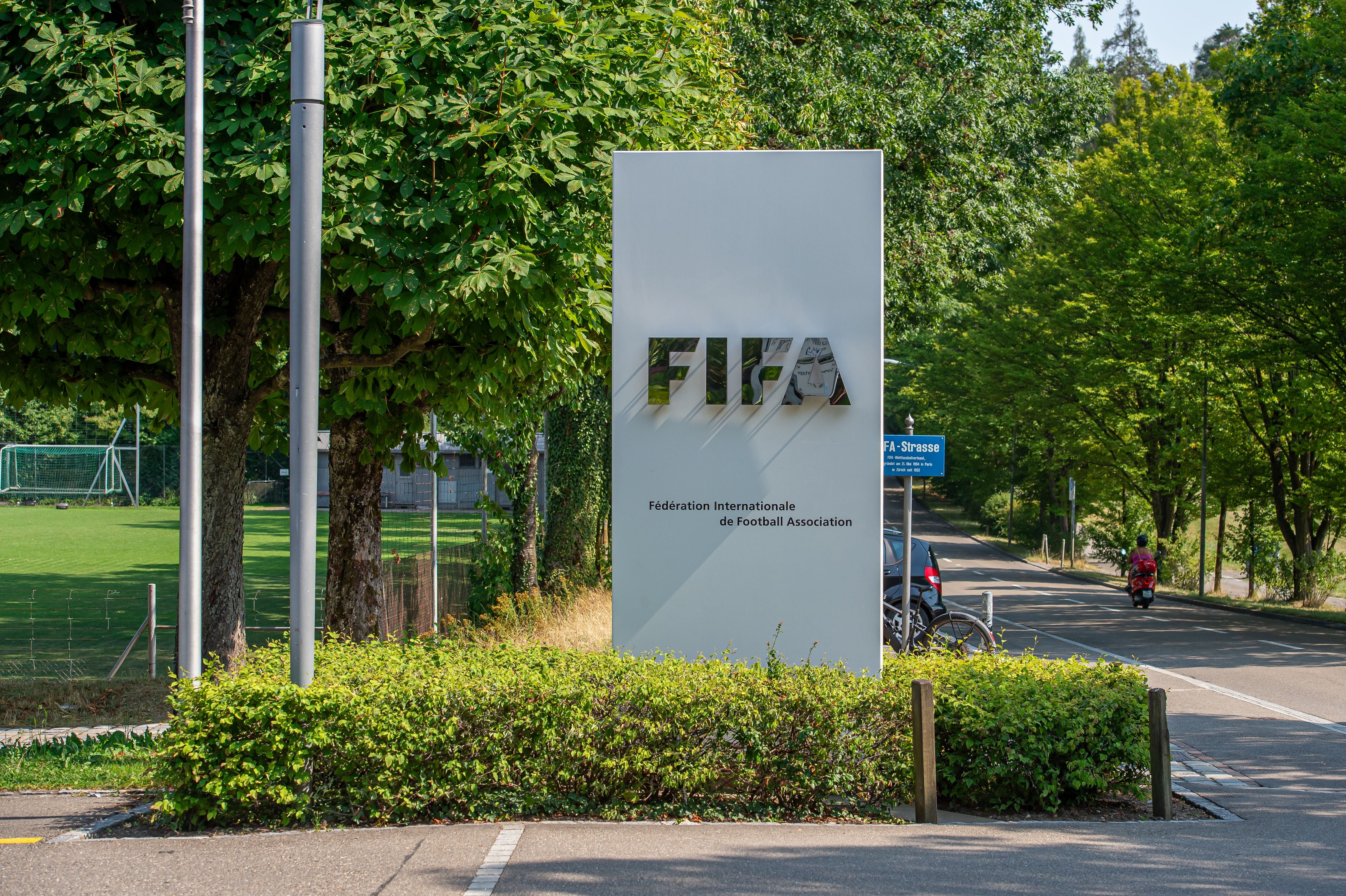 Eingangsschild der FIFA-Zentrale in Zürich vor grünem Fußballfeld und Bäumen.