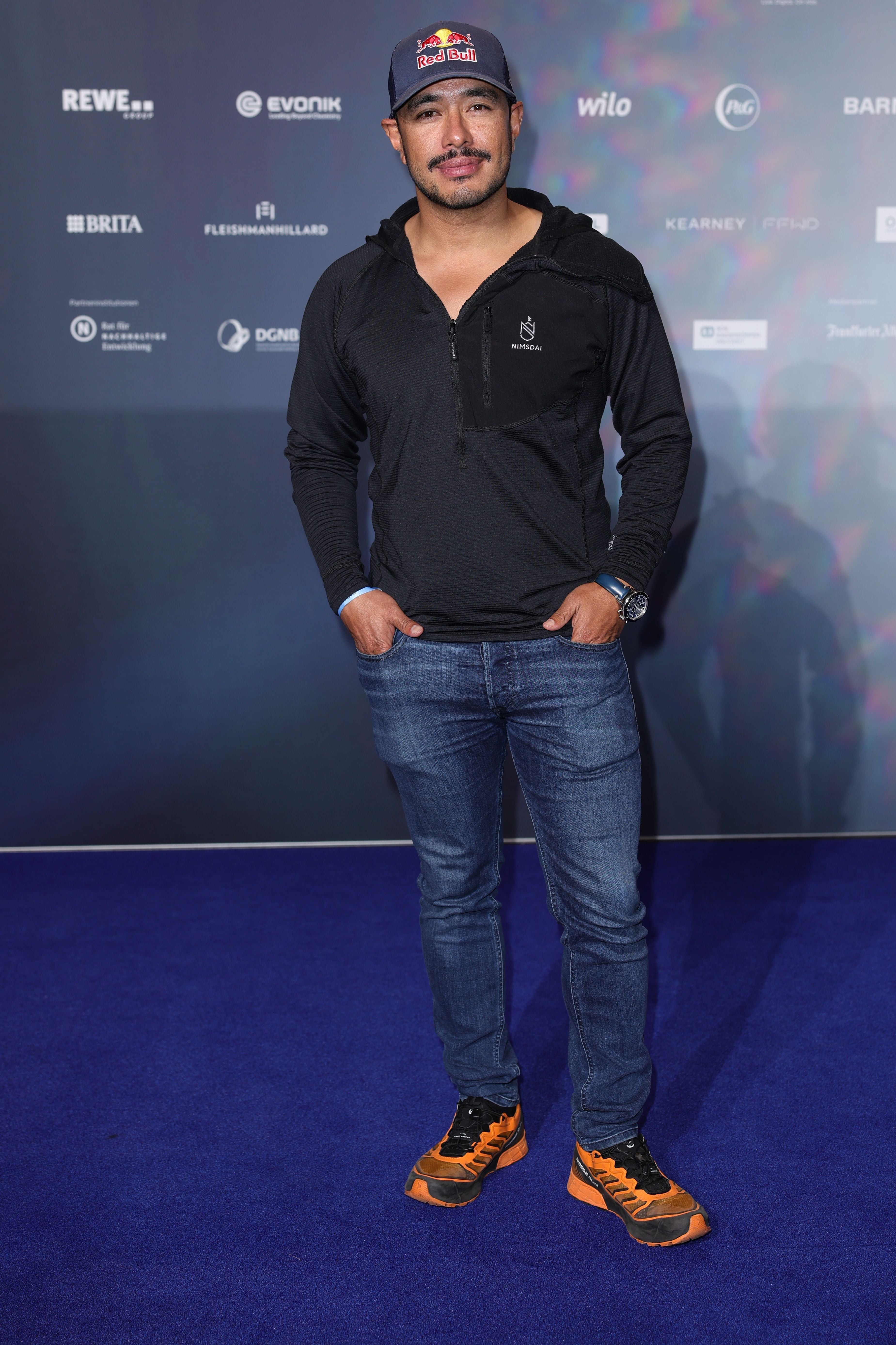 Ein Mann mit schwarzem Hoodie, Jeans und Kappe steht auf blauem Teppich vor Fotowand.