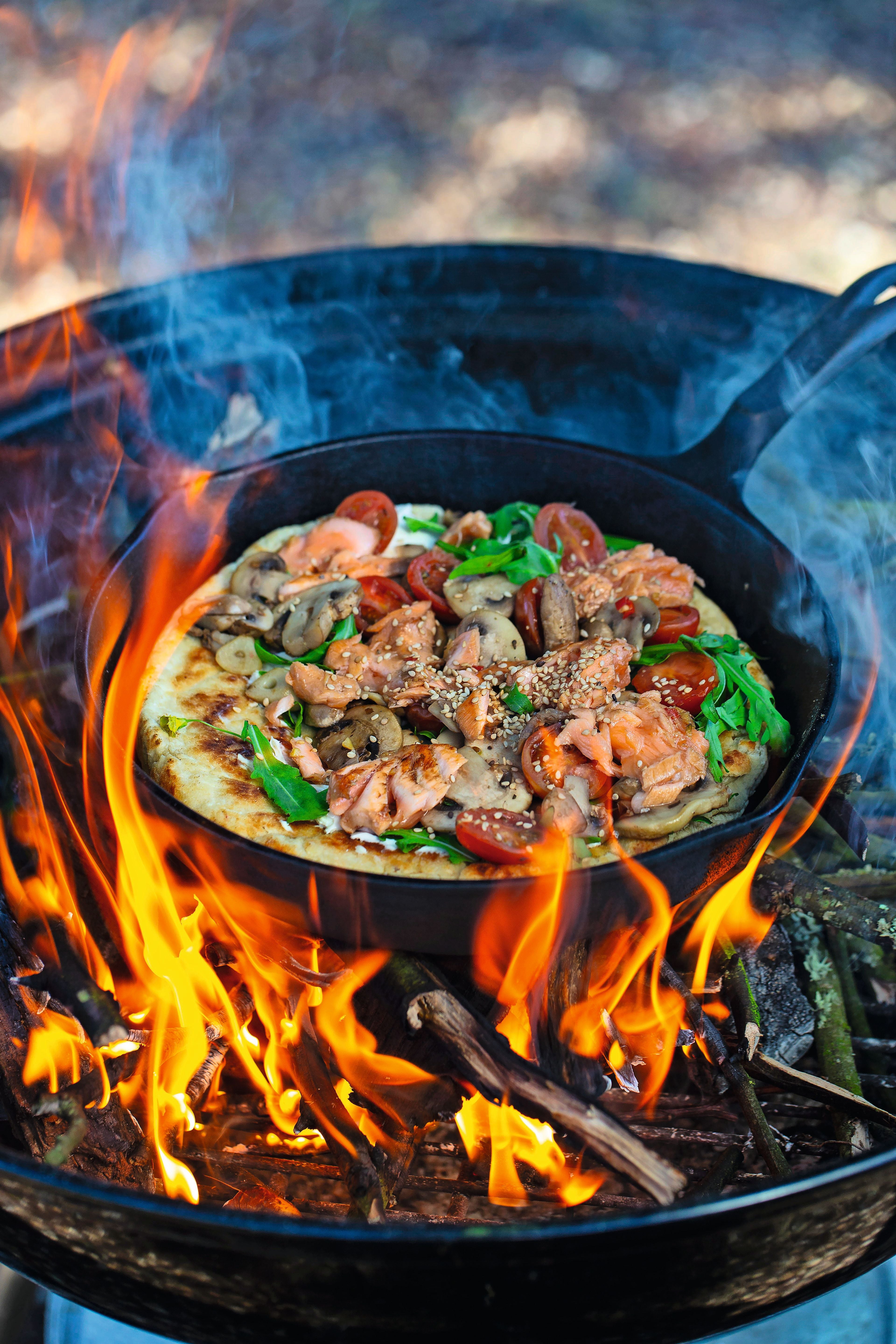 Pfannenpizza mit Lachs, Tomaten und Gemüse wird über offenem Grillfeuer zubereitet.