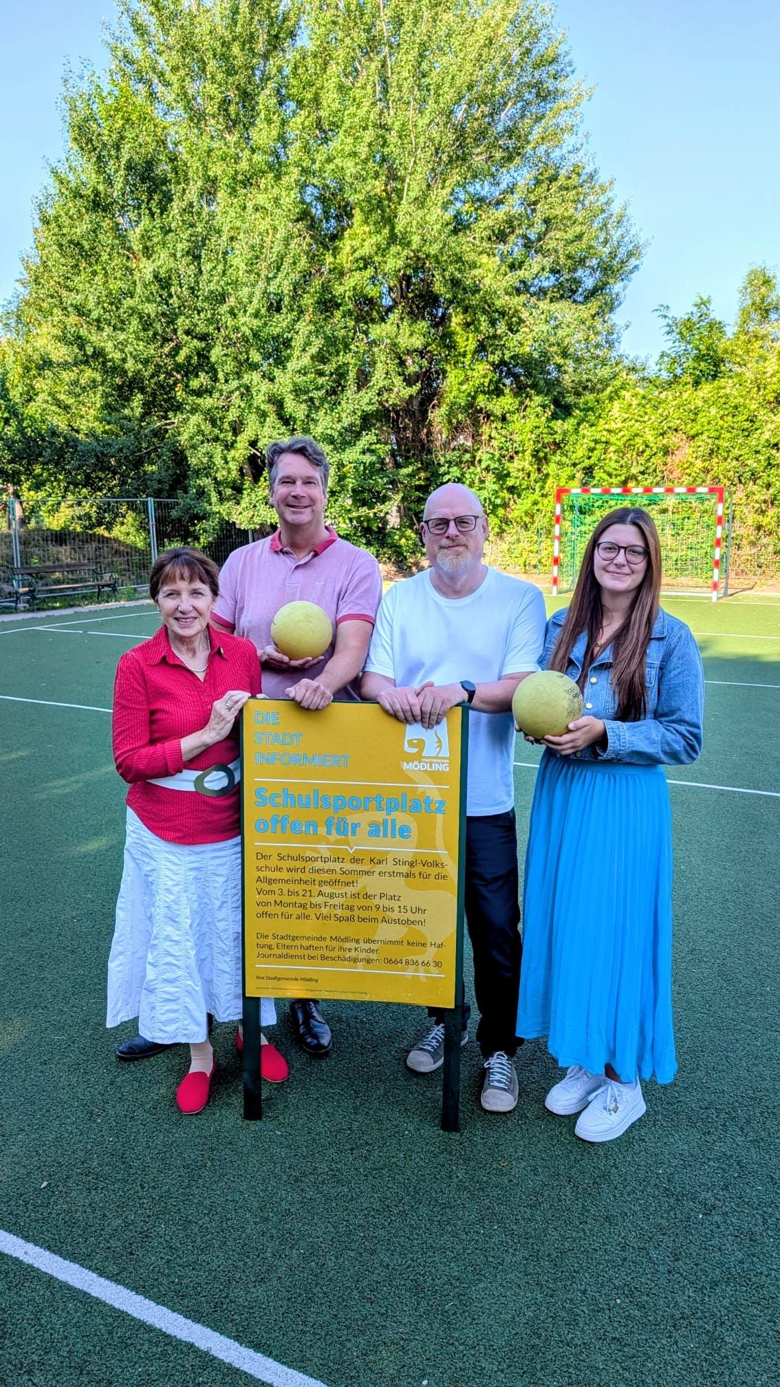 Vier Personen stehen auf einem Sportplatz neben einem gelben Hinweisschild und halten Bälle.