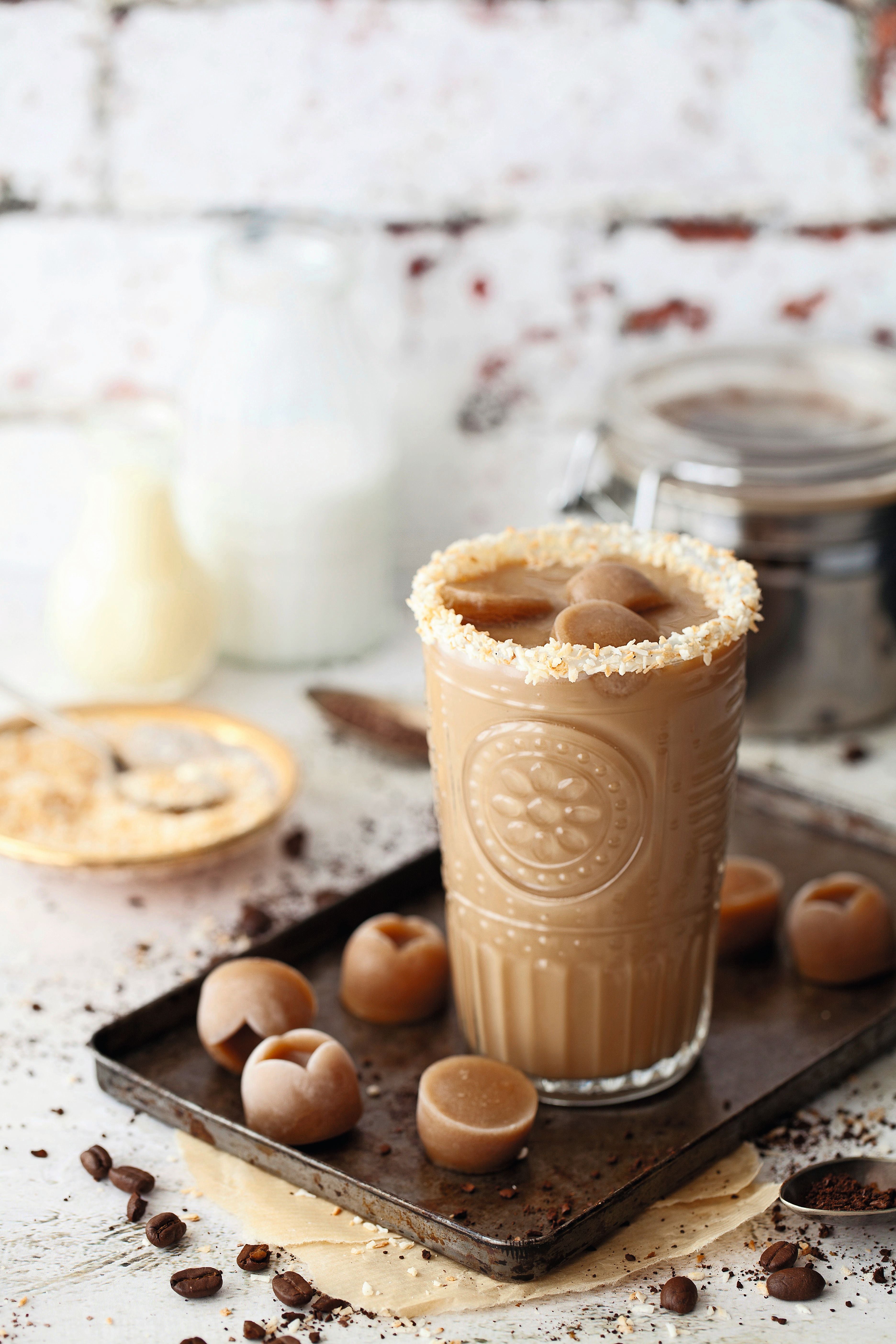 Glas Eiskaffee mit Kaffeeeiswürfeln und Kokosrand auf einem Tablett, umgeben von Kaffeebohnen.