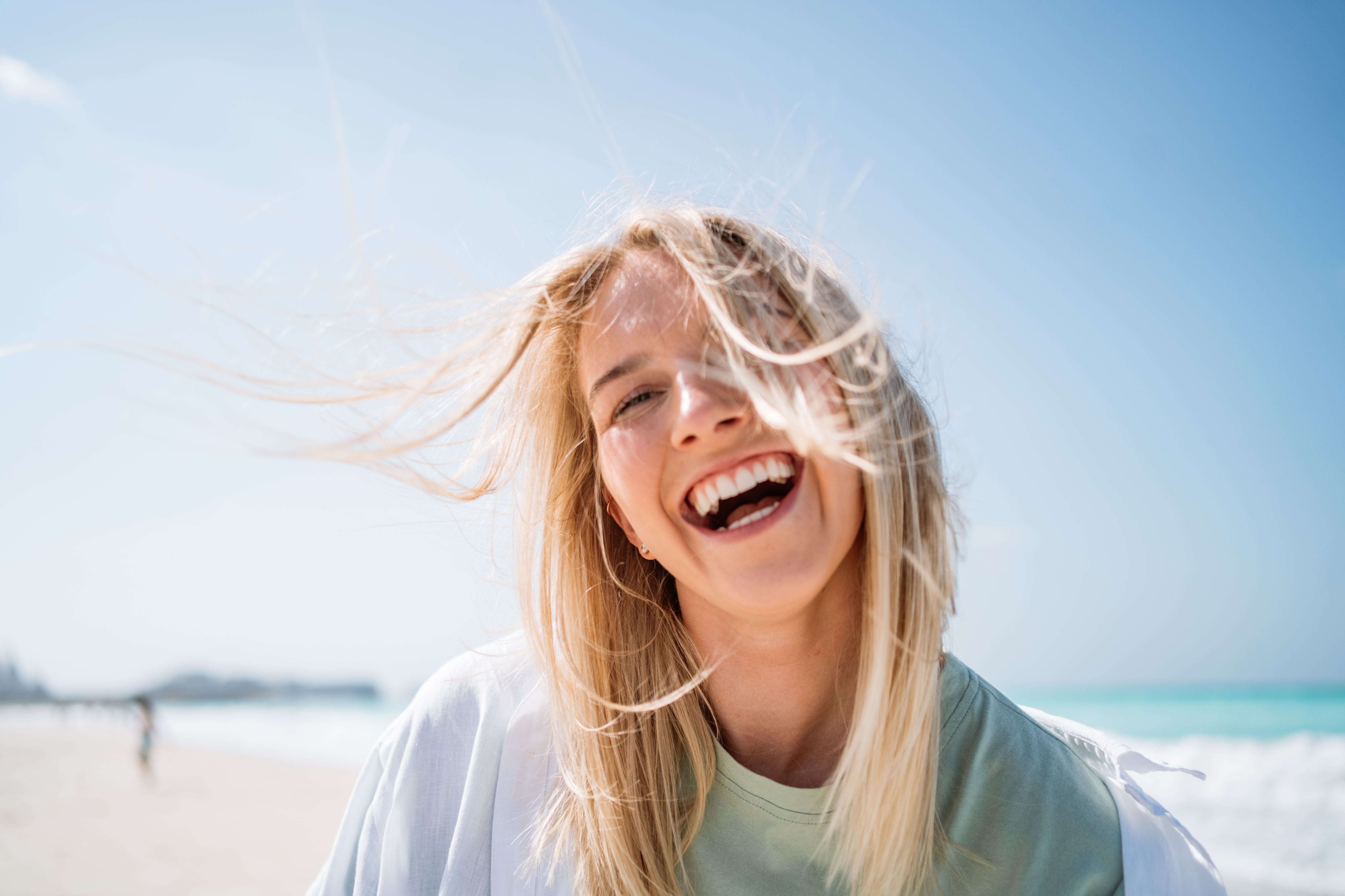 Lachende Frau mit wehendem Haar am Strand vor blauem Himmel.