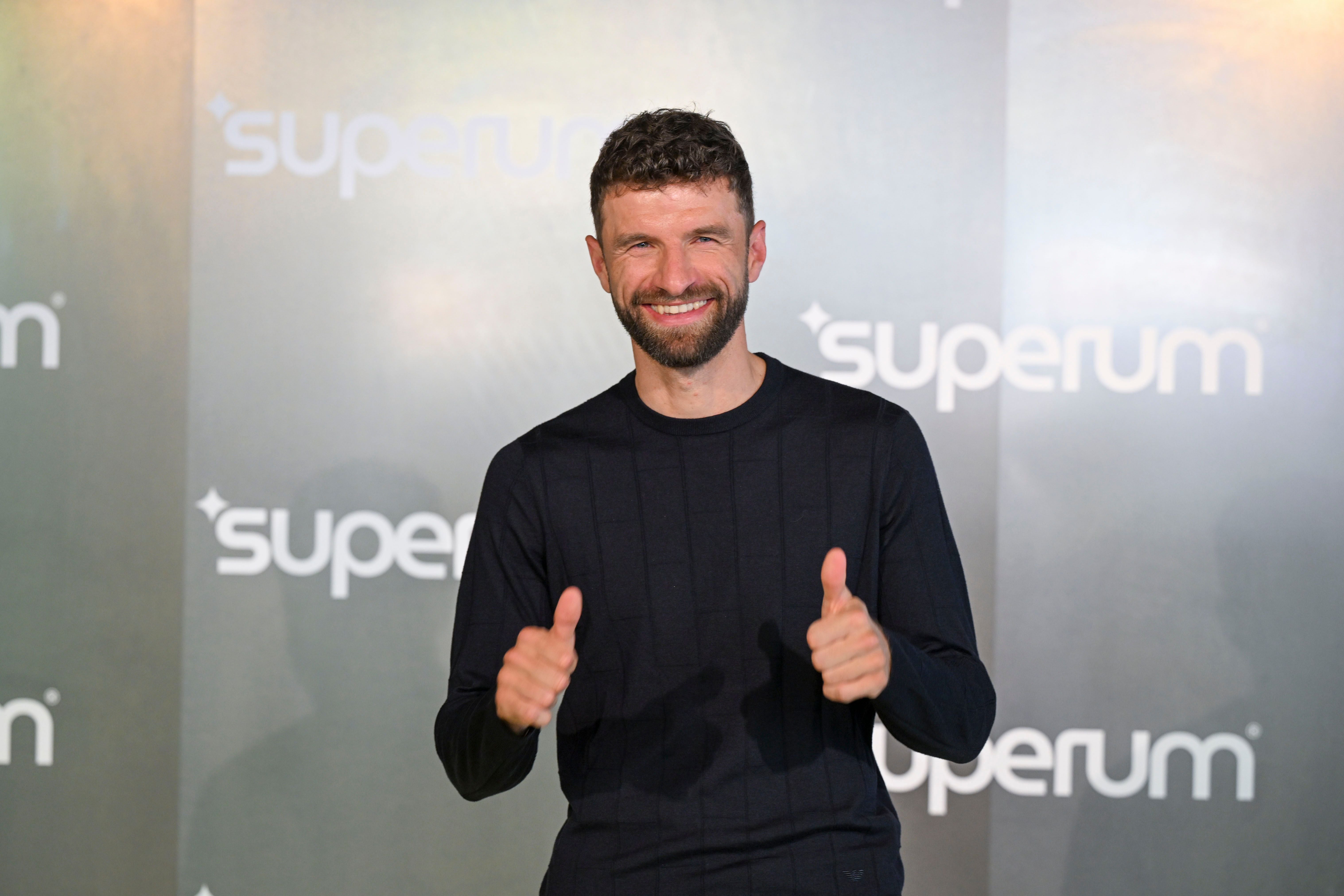 Ein Mann mit Bart lächelt und zeigt beide Daumen hoch vor einer "superum"-Wand.