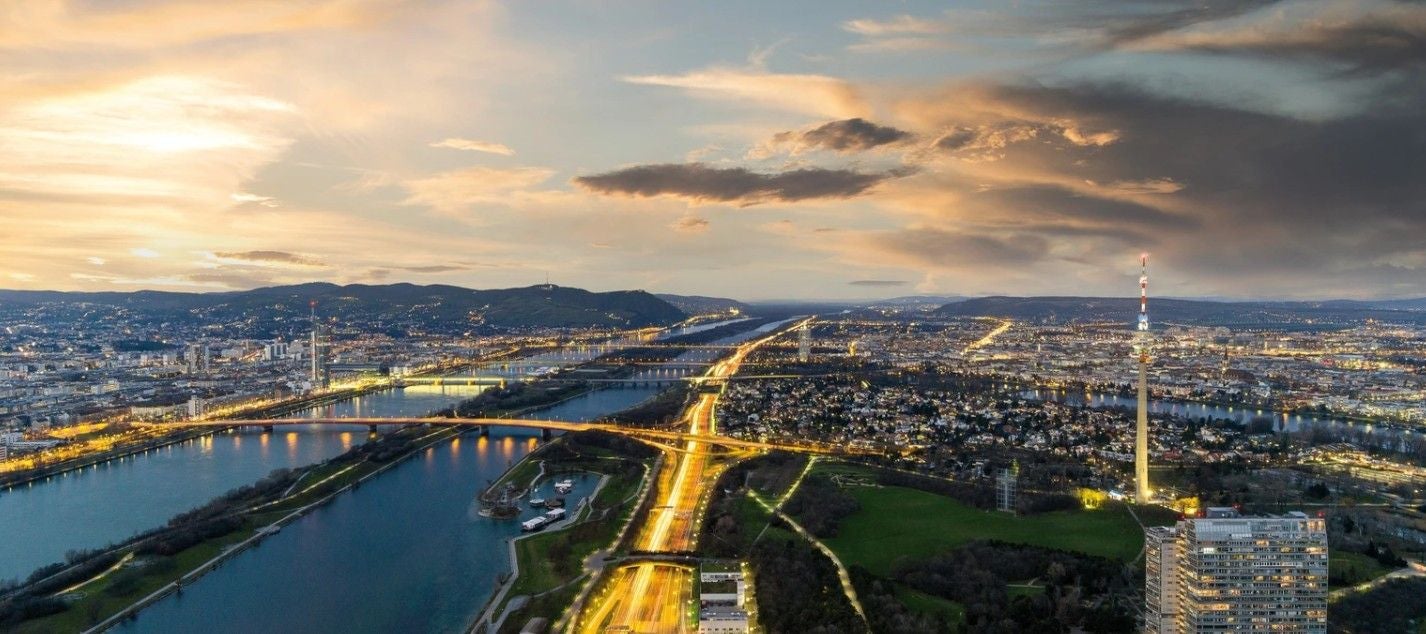 Blick auf Wien bei Sonnenuntergang mit der Donau, beleuchteten Straßen und dem Donauturm.