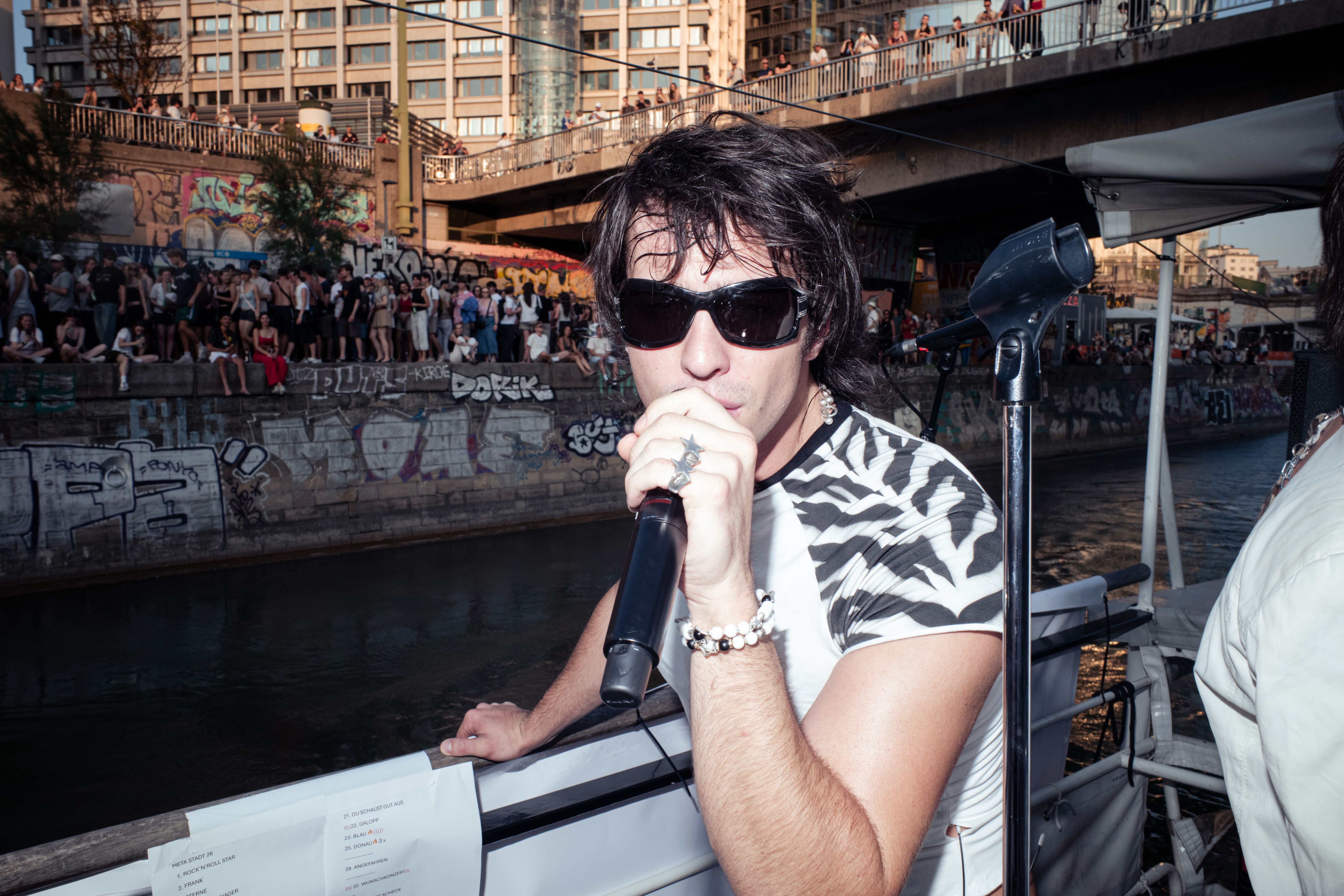 Ein Sänger mit Sonnenbrille performt am Fluss vor einer graffiti-besprühten Mauer und Publikum.