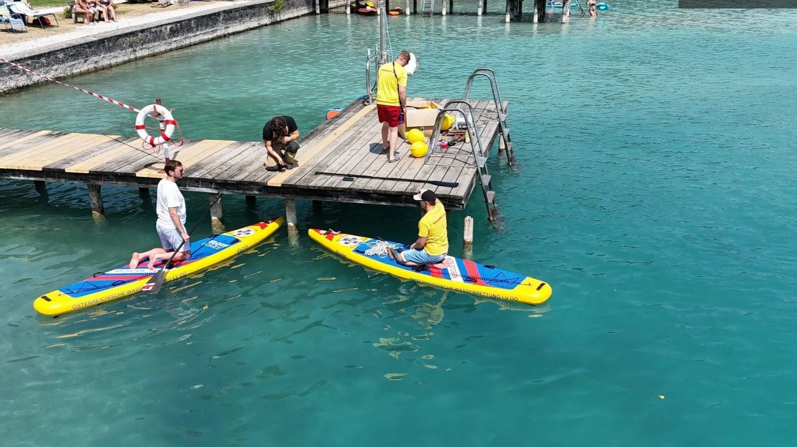 Zwei Rettungskräfte auf gelben SUP-Boards neben einem Steg im klaren Wasser, weitere Personen am Steg.