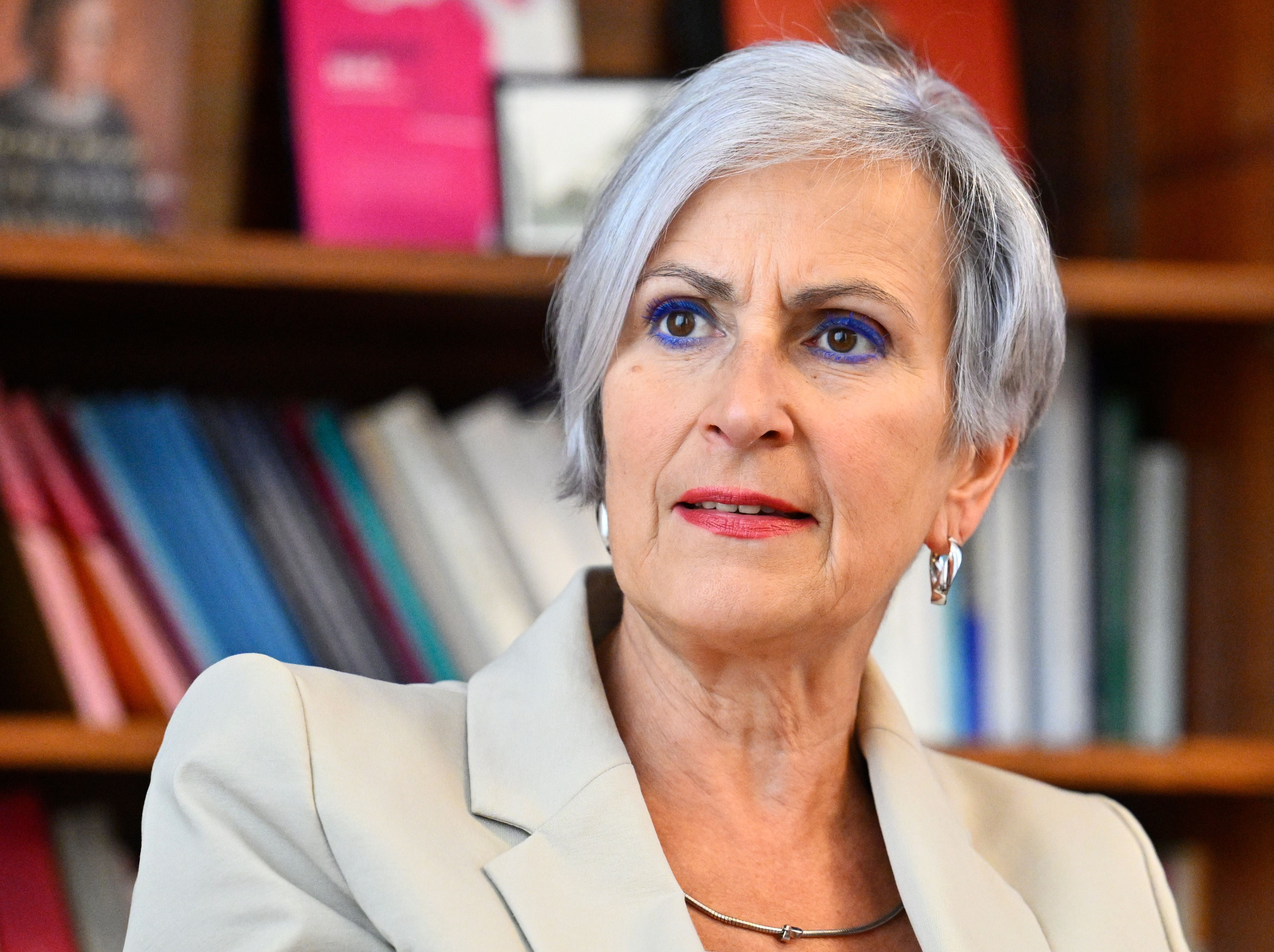 Frau mit grauen Haaren und hellem Blazer sitzt vor einem Bücherregal.