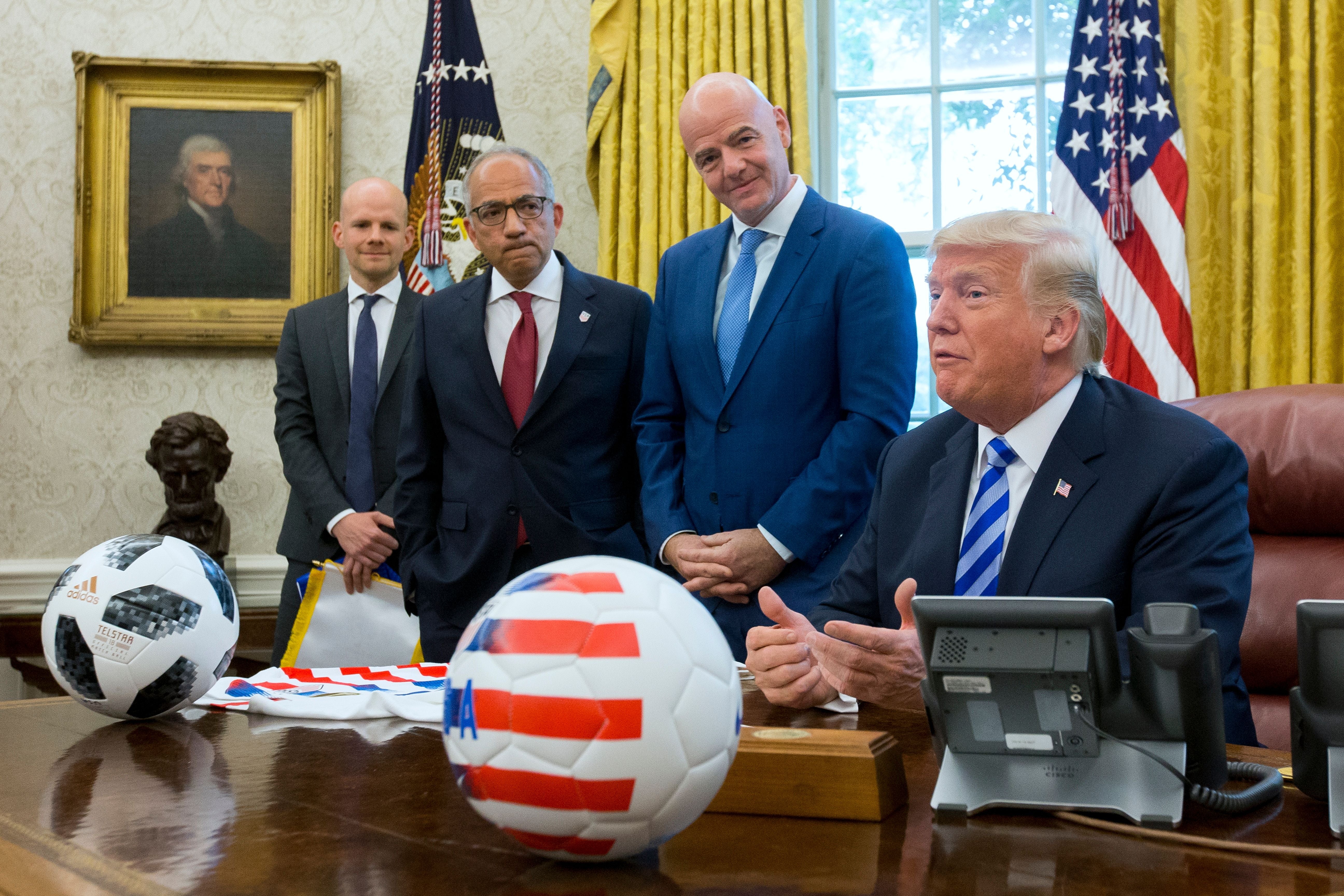 Vier Männer im Anzug mit zwei Fußballbällen im Oval Office des Weißen Hauses, US-Flagge im Hintergrund.