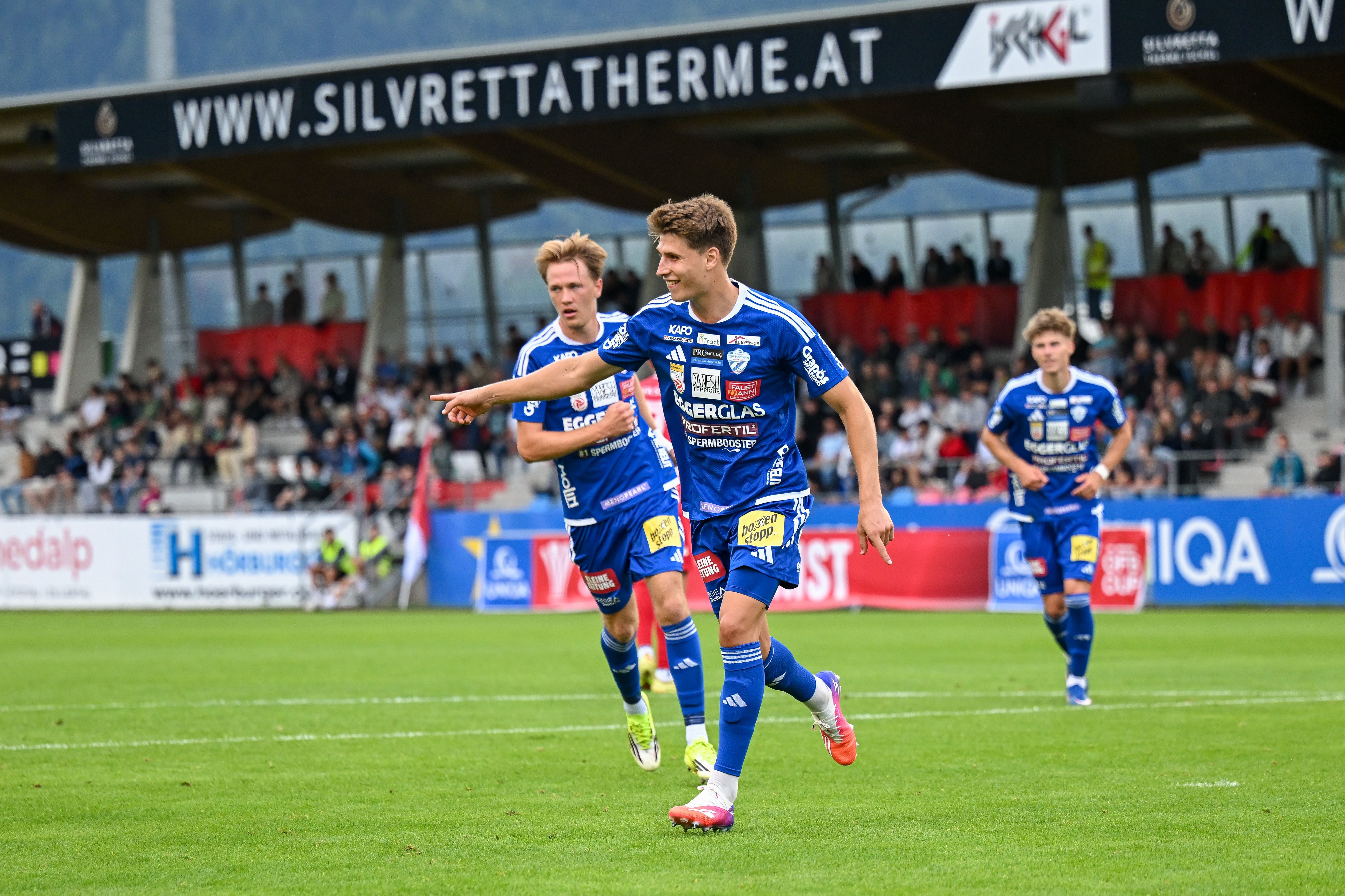 Drei Fußballspieler im blauen Trikot jubeln auf dem Spielfeld vor Zuschauern.
