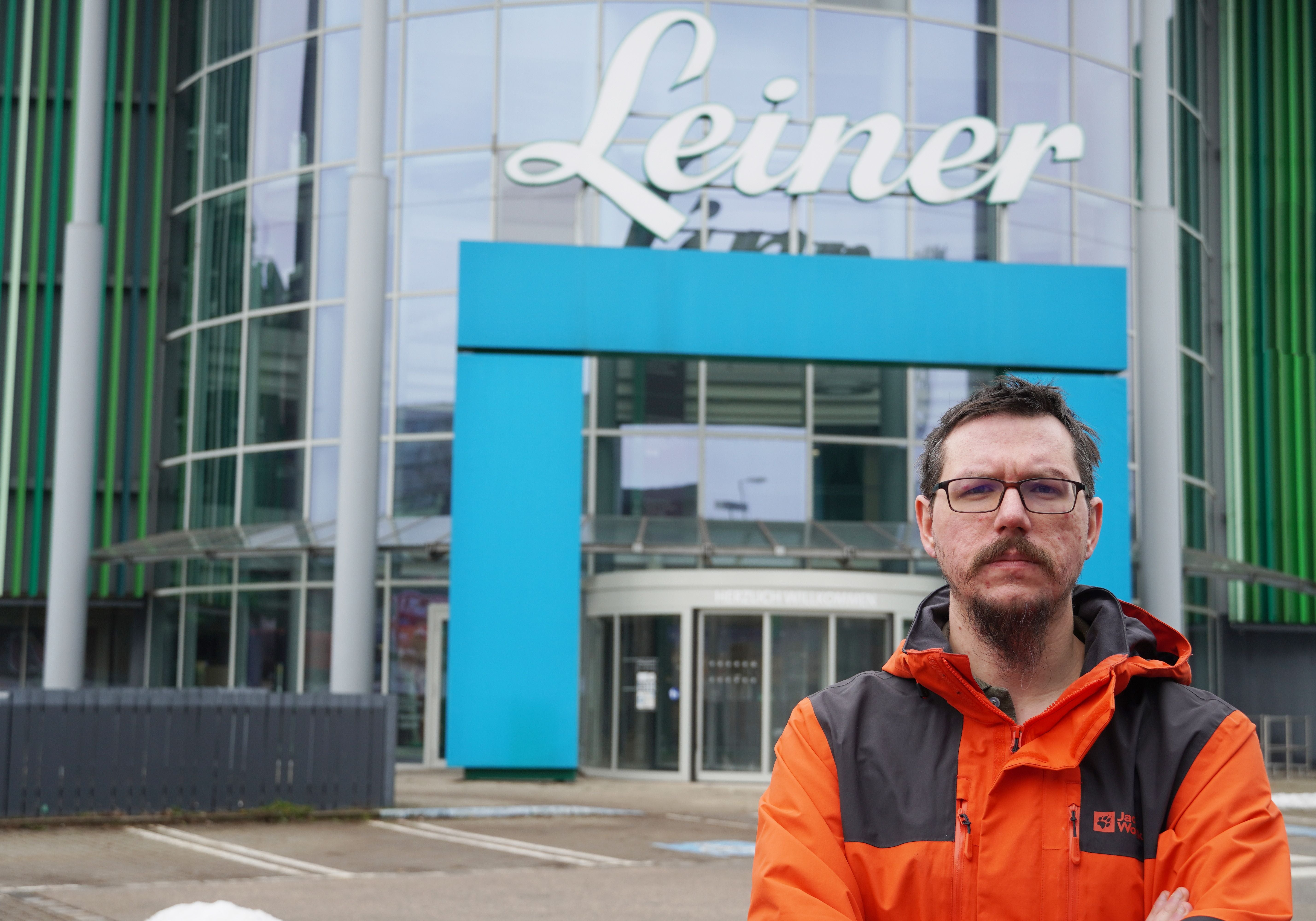 Ein Mann mit Brille und orangener Jacke steht vor einem Gebäude mit dem Schriftzug "Leiner".