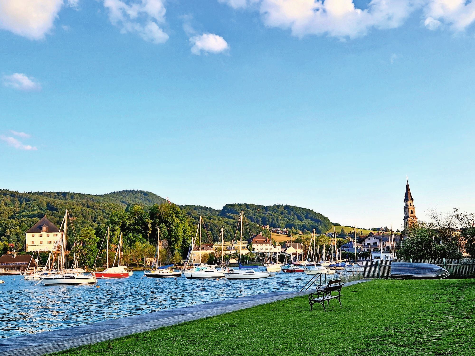 Segelboote liegen am Ufer des Mattsees, mit grünen Hügeln und Dorf im Hintergrund.