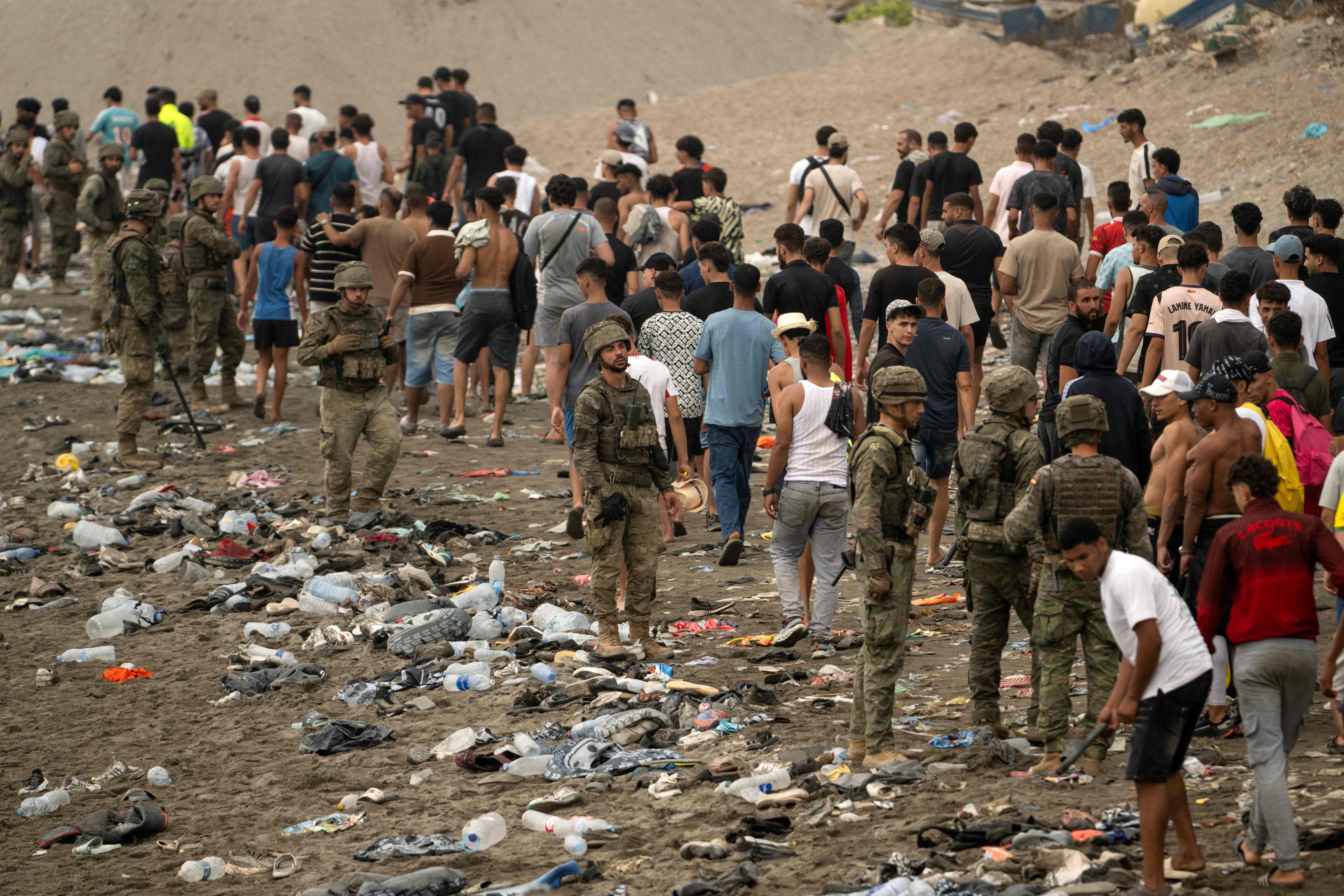 Soldaten überwachen eine große Gruppe von Migranten an einem verschmutzten Strand.