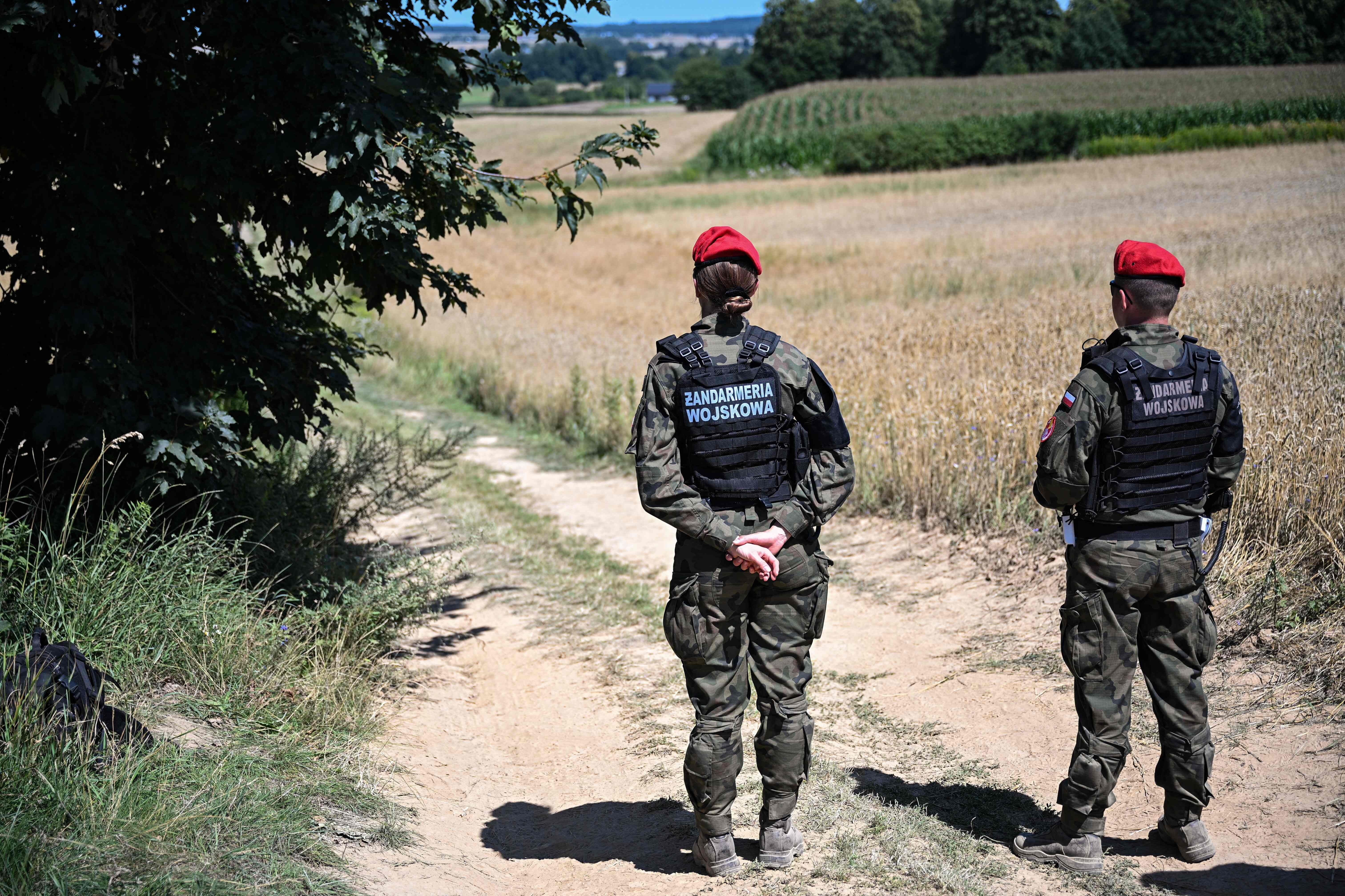 Zwei Militärgendarmeristen mit roten Baretten bewachen einen Feldweg am Rand eines Kornfelds.