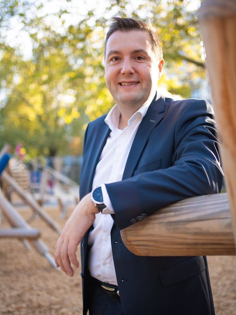 Ein Mann im Anzug lehnt lächelnd an einem Holzgeländer im Freien bei Sonnenschein.