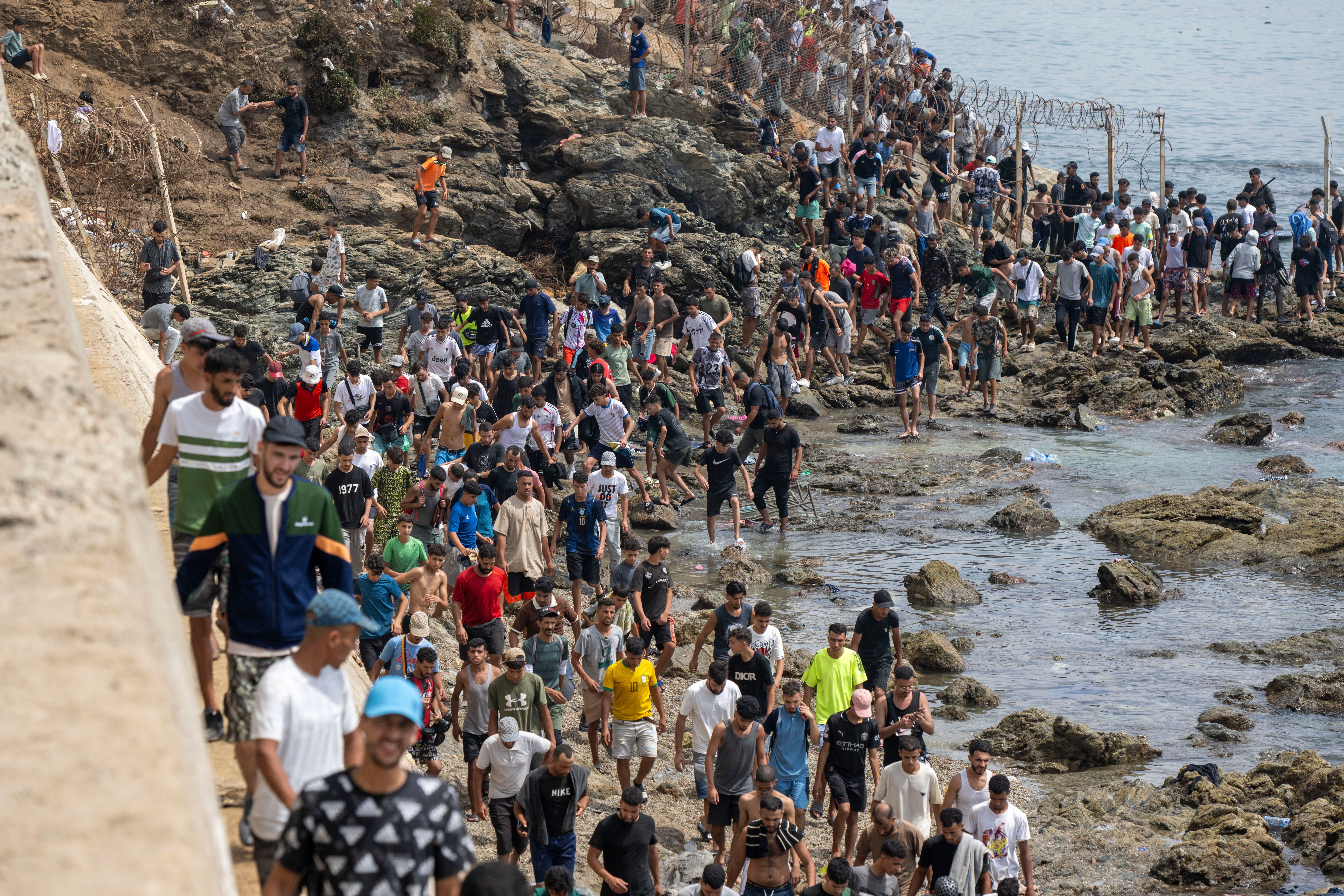 Viele junge Männer versammeln sich an einem felsigen Küstenabschnitt nahe einem Grenzzaun.