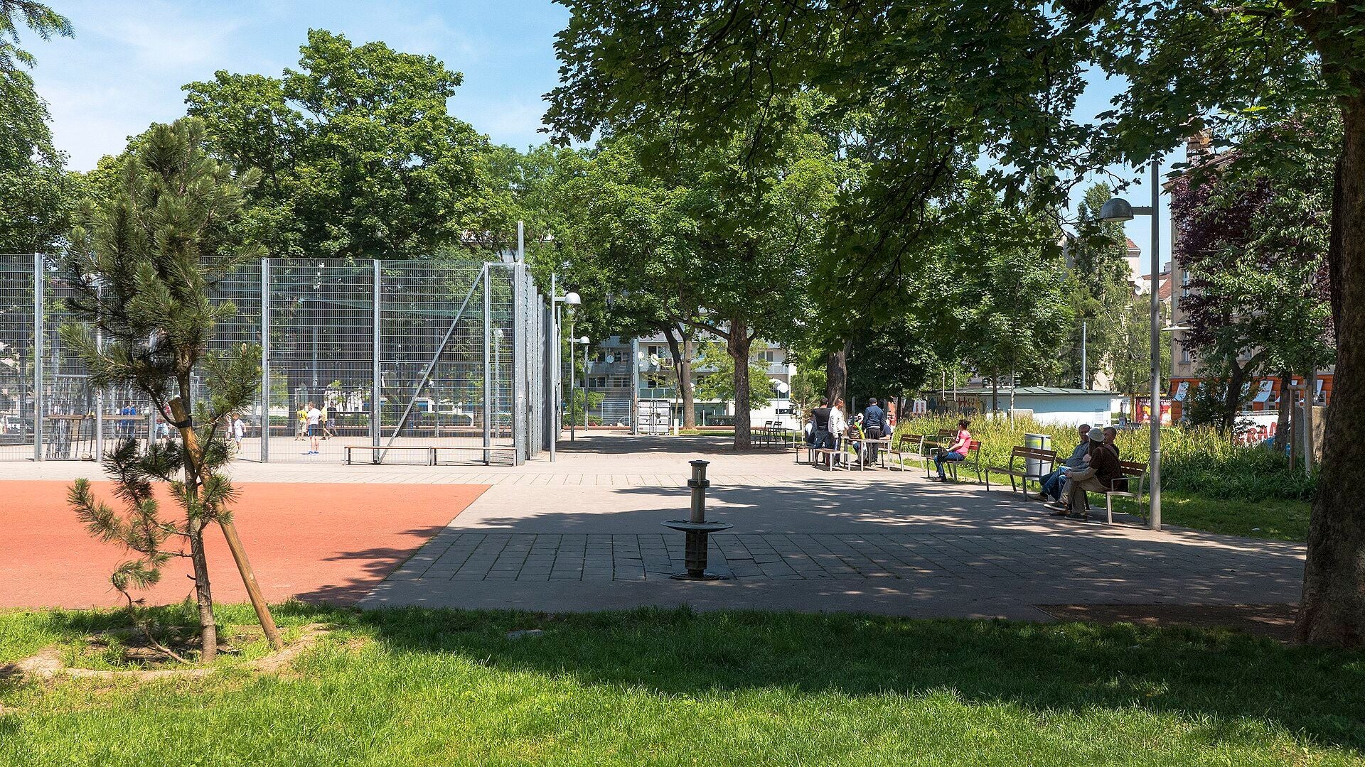 Mehrere Personen sitzen auf Parkbänken im Schatten, andere spielen auf einem Sportplatz.