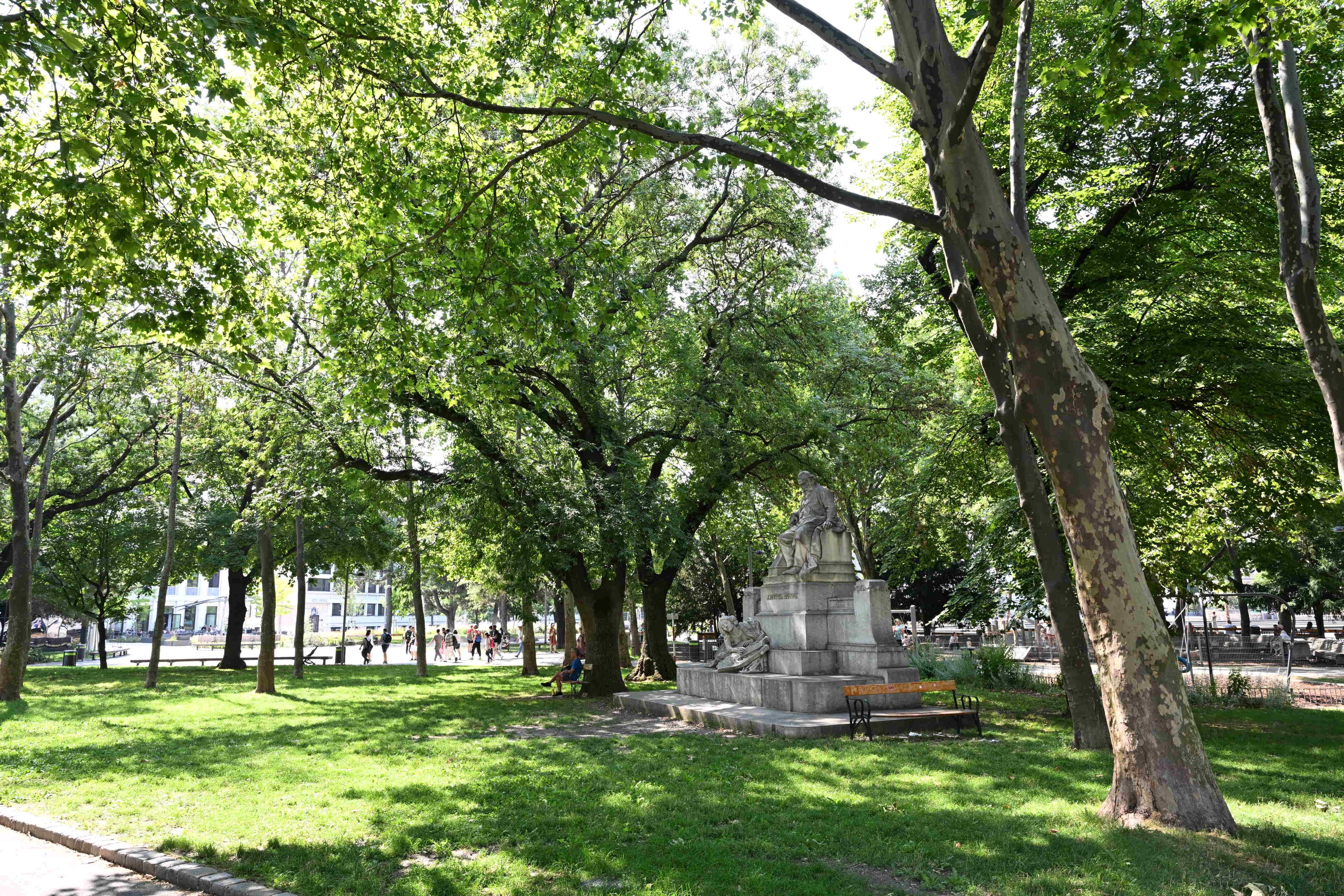 Grüner Park am Karlsplatz in Wien mit Bäumen und einer Statue im Sonnenschein.