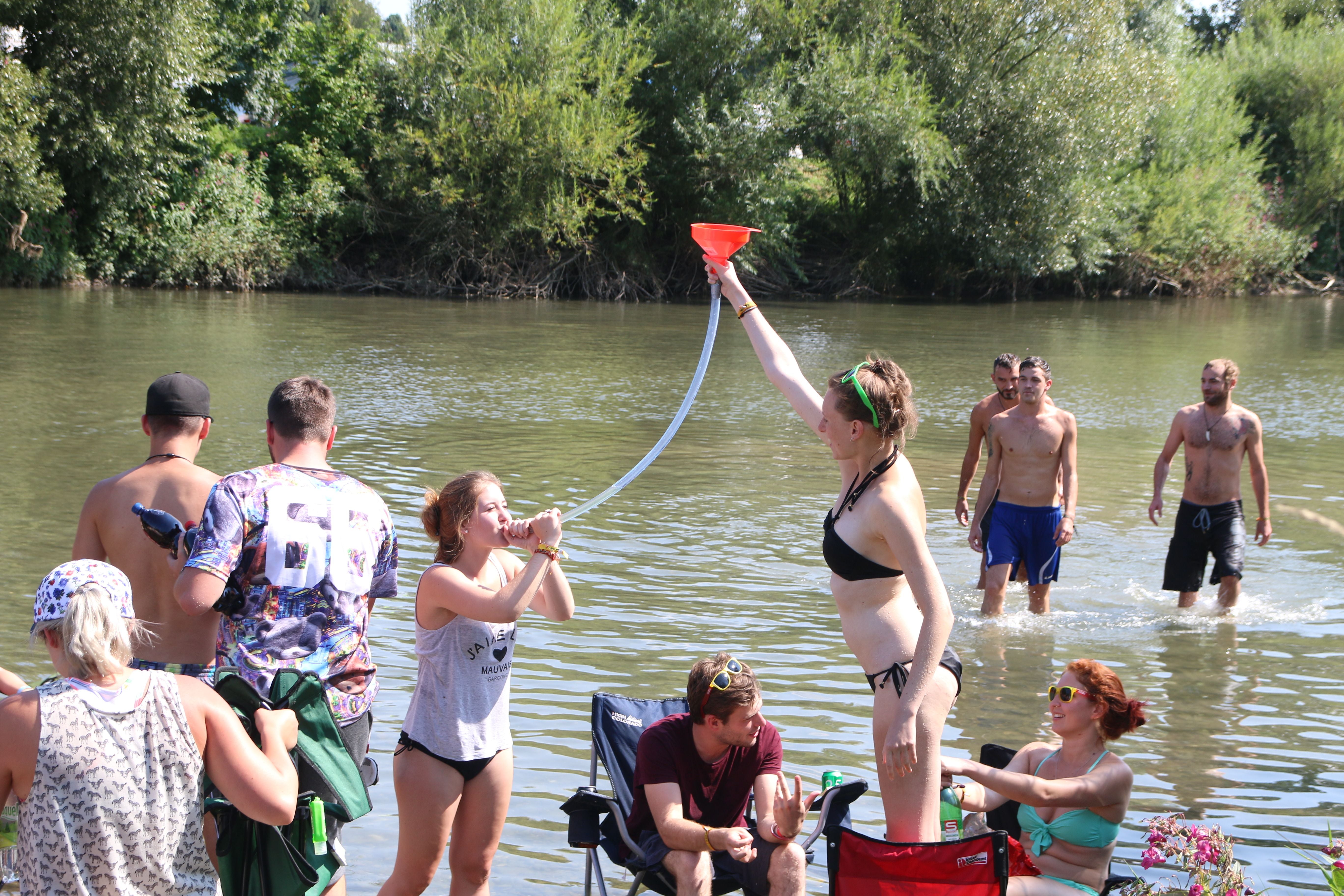 Junge Leute feiern im Wasser, eine Frau nutzt einen Biertrichter, andere entspannen am Ufer.