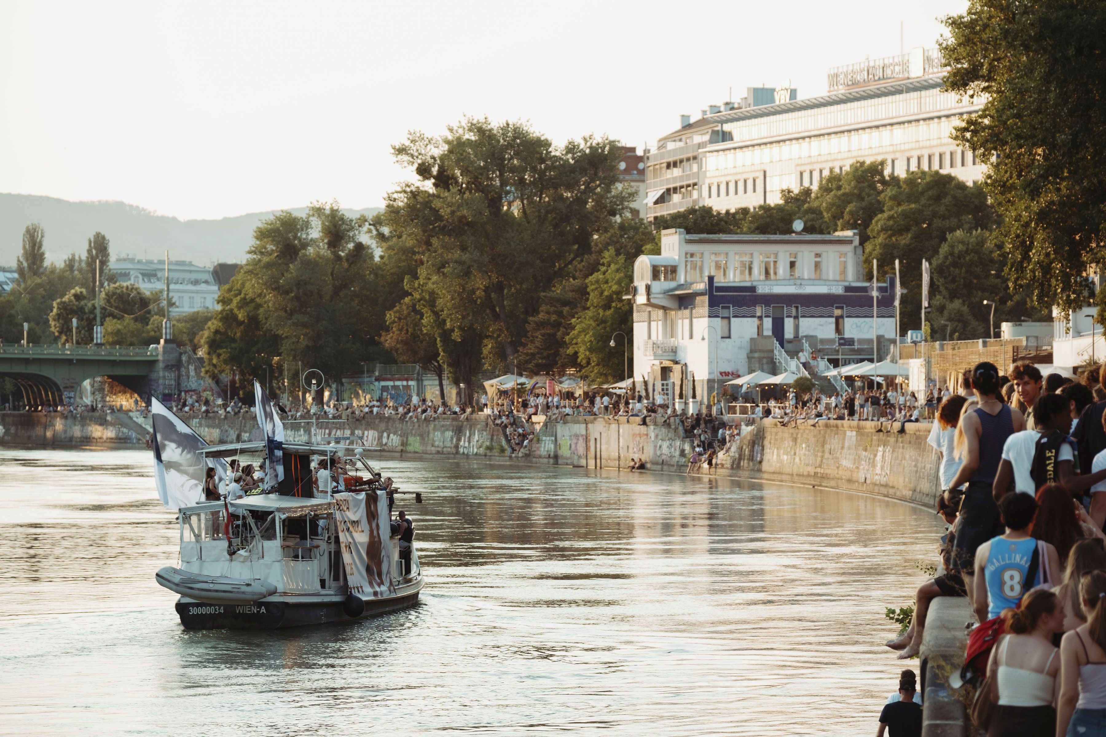 Boot mit Menschen auf einem Fluss, viele Leute sitzen am sonnigen Ufer in einer Stadt.
