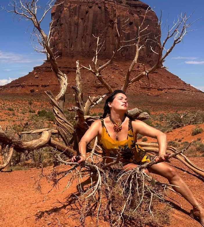 Frau posiert in gelbem Kleid vor karger Landschaft mit Felsen und trockenem Baum im Hintergrund.
