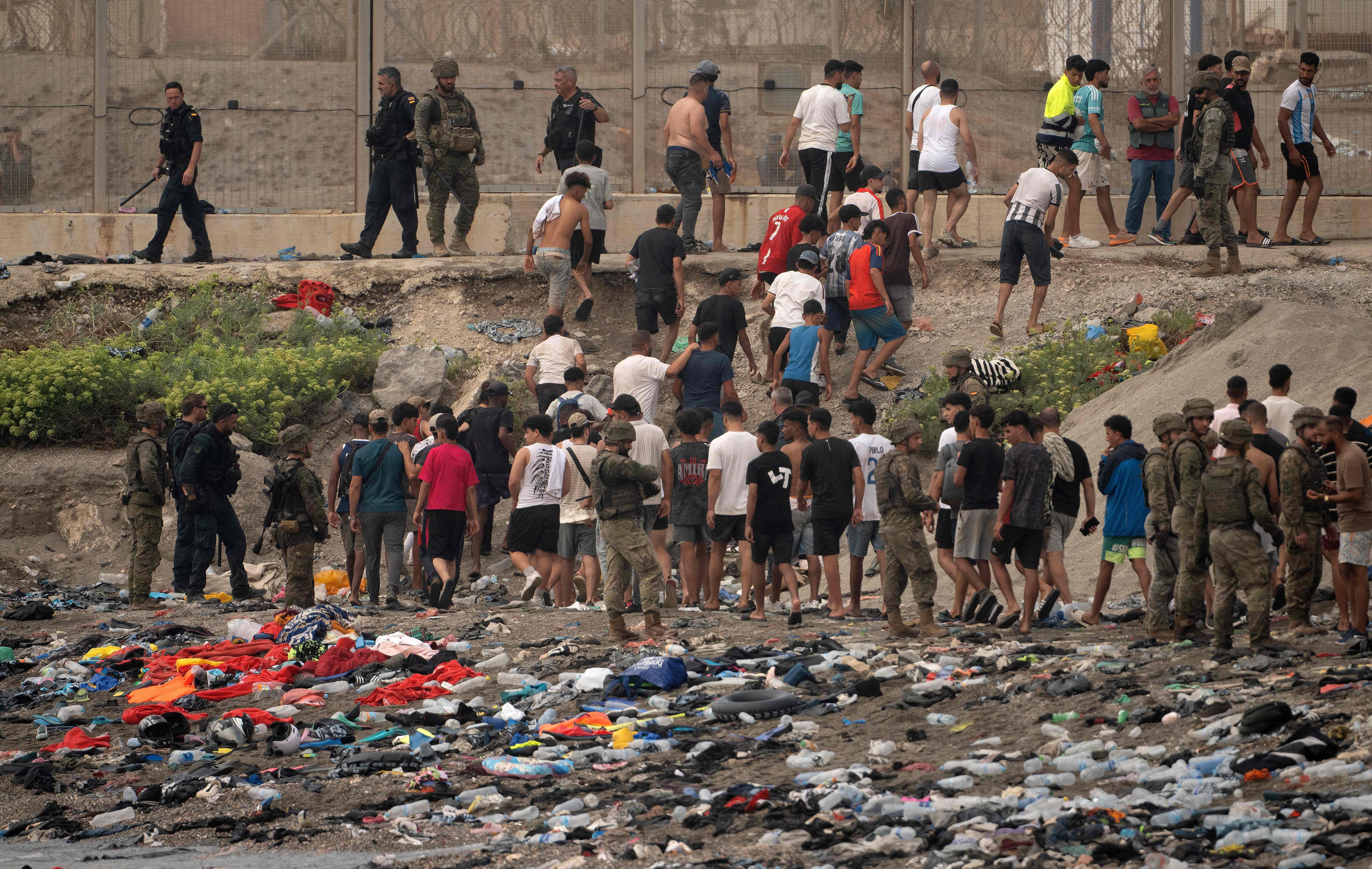 Soldaten und Polizisten kontrollieren eine Gruppe Migranten an einem Strand voller zurückgelassener Kleidung.