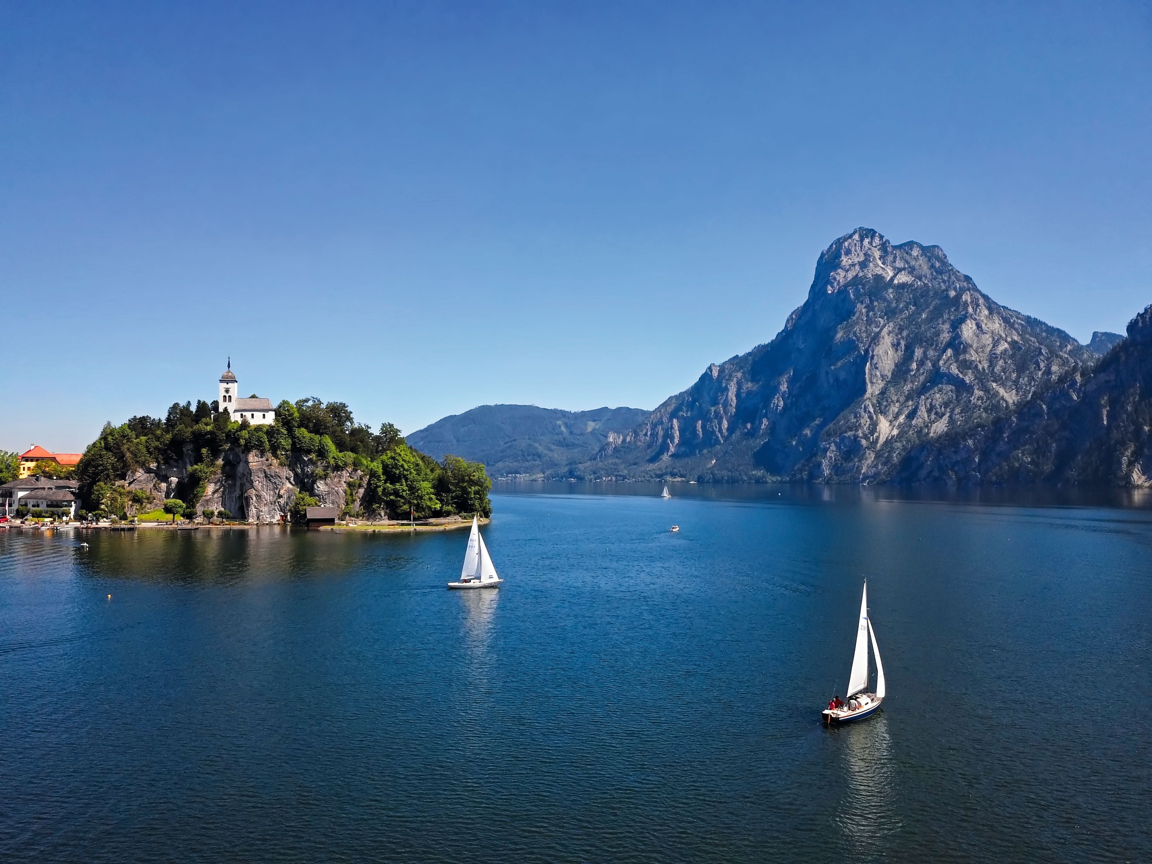 Blick auf die Traunkirche am See mit Segelbooten und Bergen im Hintergrund.