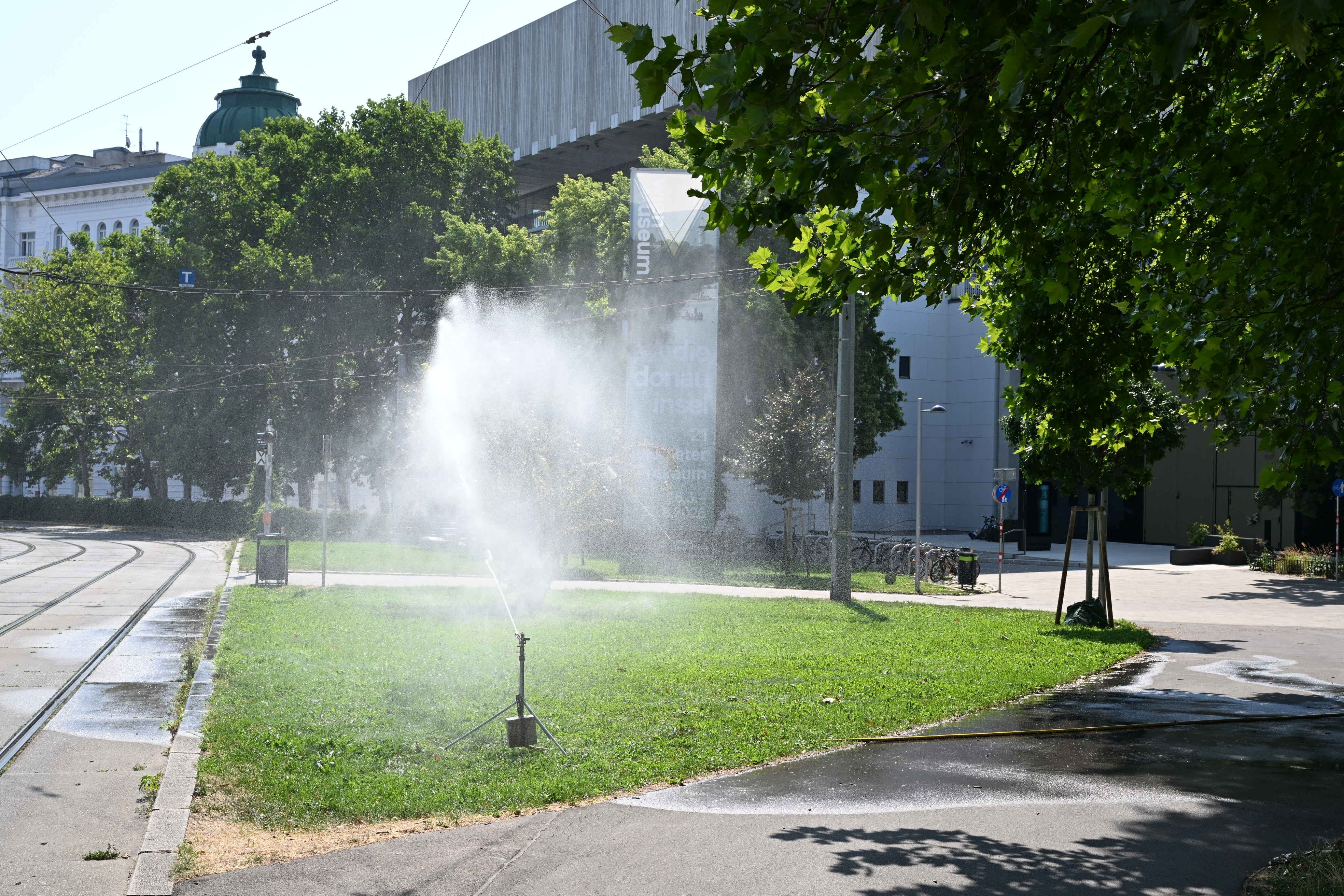 Ein Rasensprenger bewässert eine Grünfläche am Karlsplatz in Wien bei sonnigem Wetter.