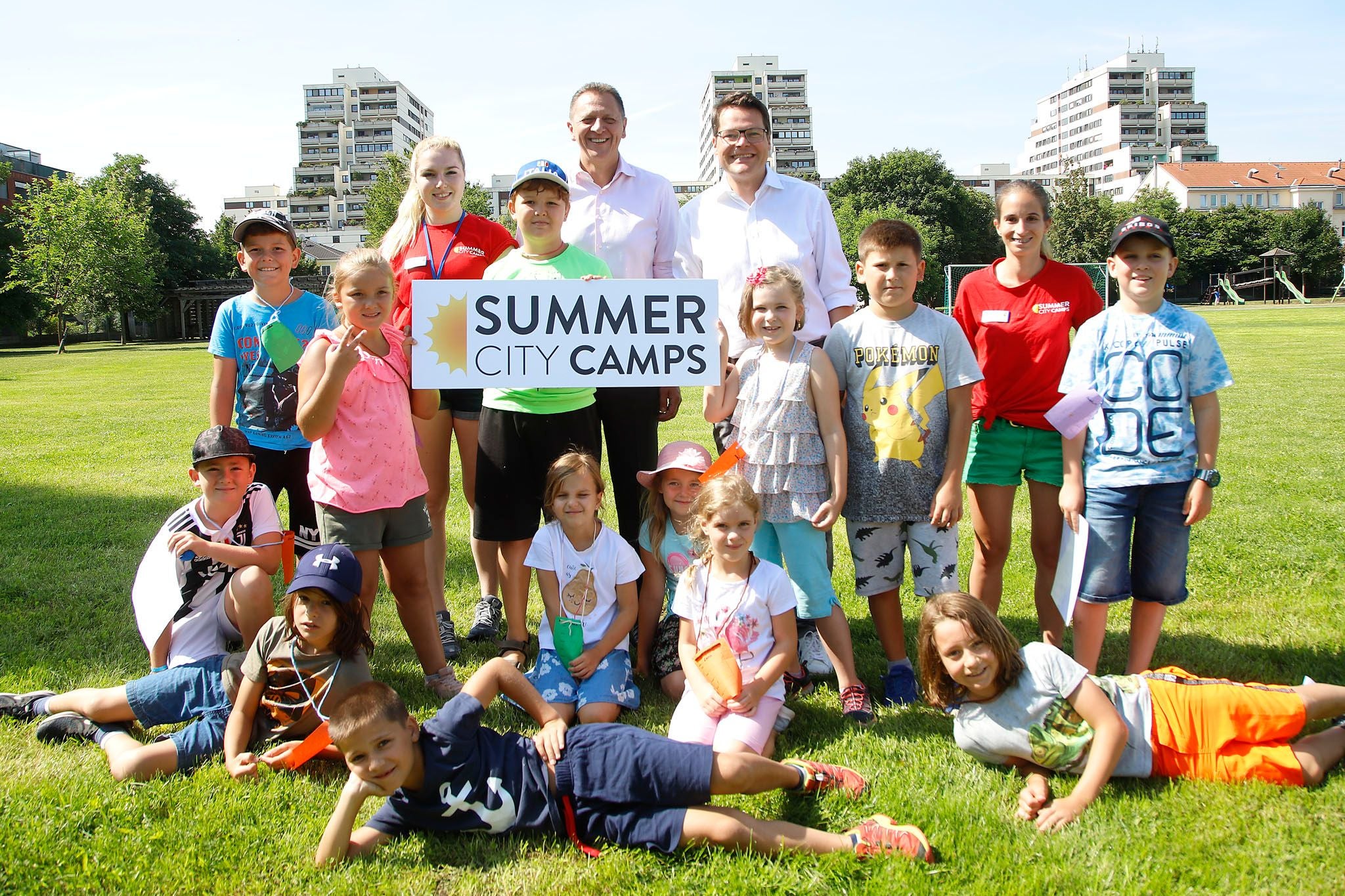 Eine Gruppe von Kindern und Erwachsenen posiert draußen mit einem „Summer City Camps“-Schild.