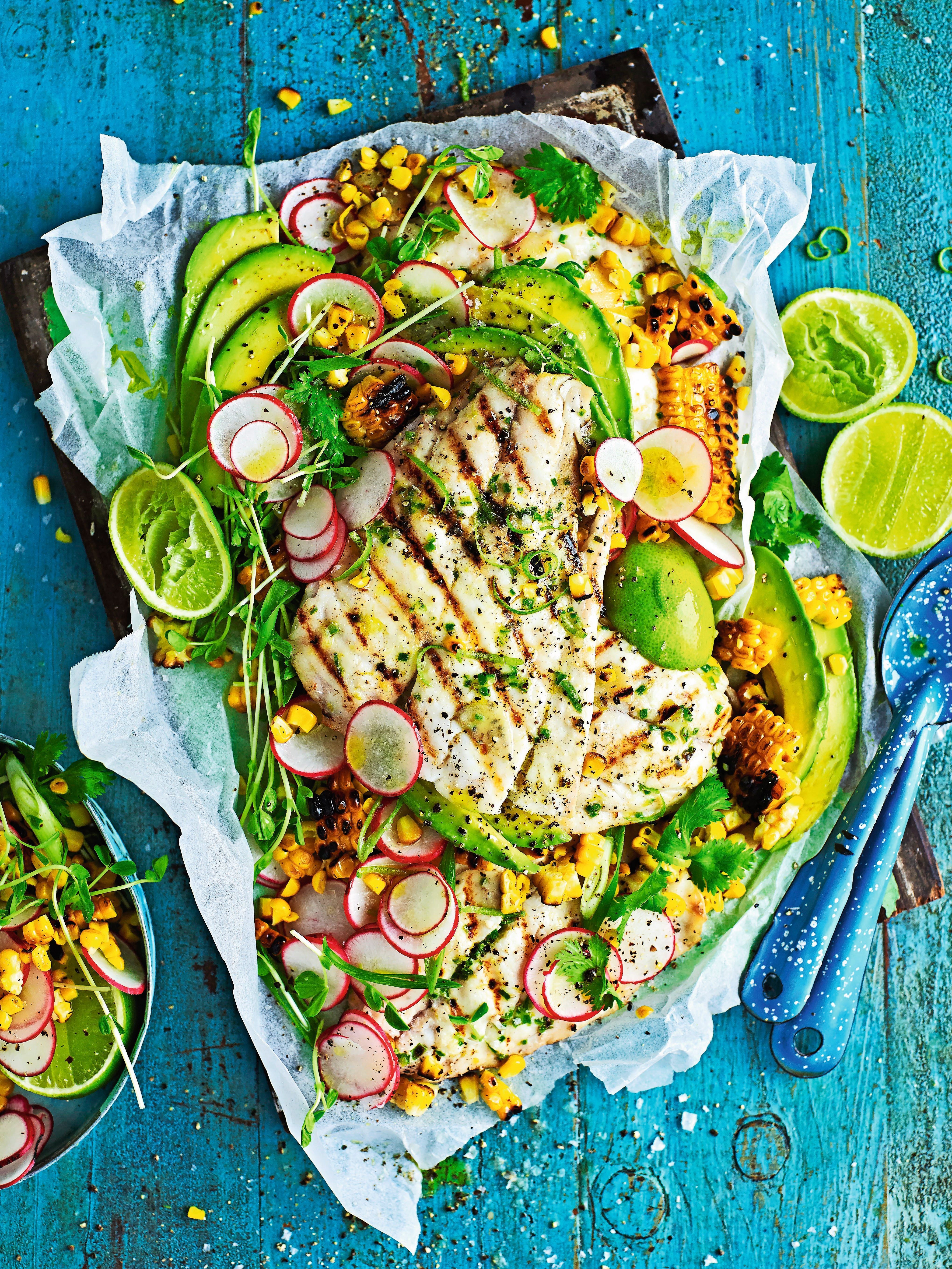 Gegrillter Fisch mit Mais-Salsa, Avocado, Radieschen und Limetten auf Backpapier angerichtet.