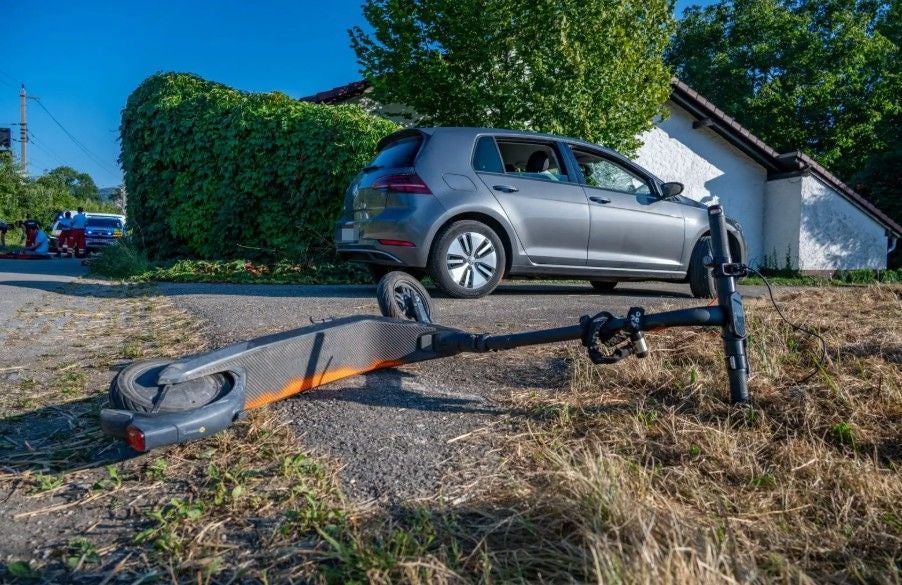 Umgestürzter E-Scooter liegt vor einem silbernen Auto auf einem ländlichen Weg.