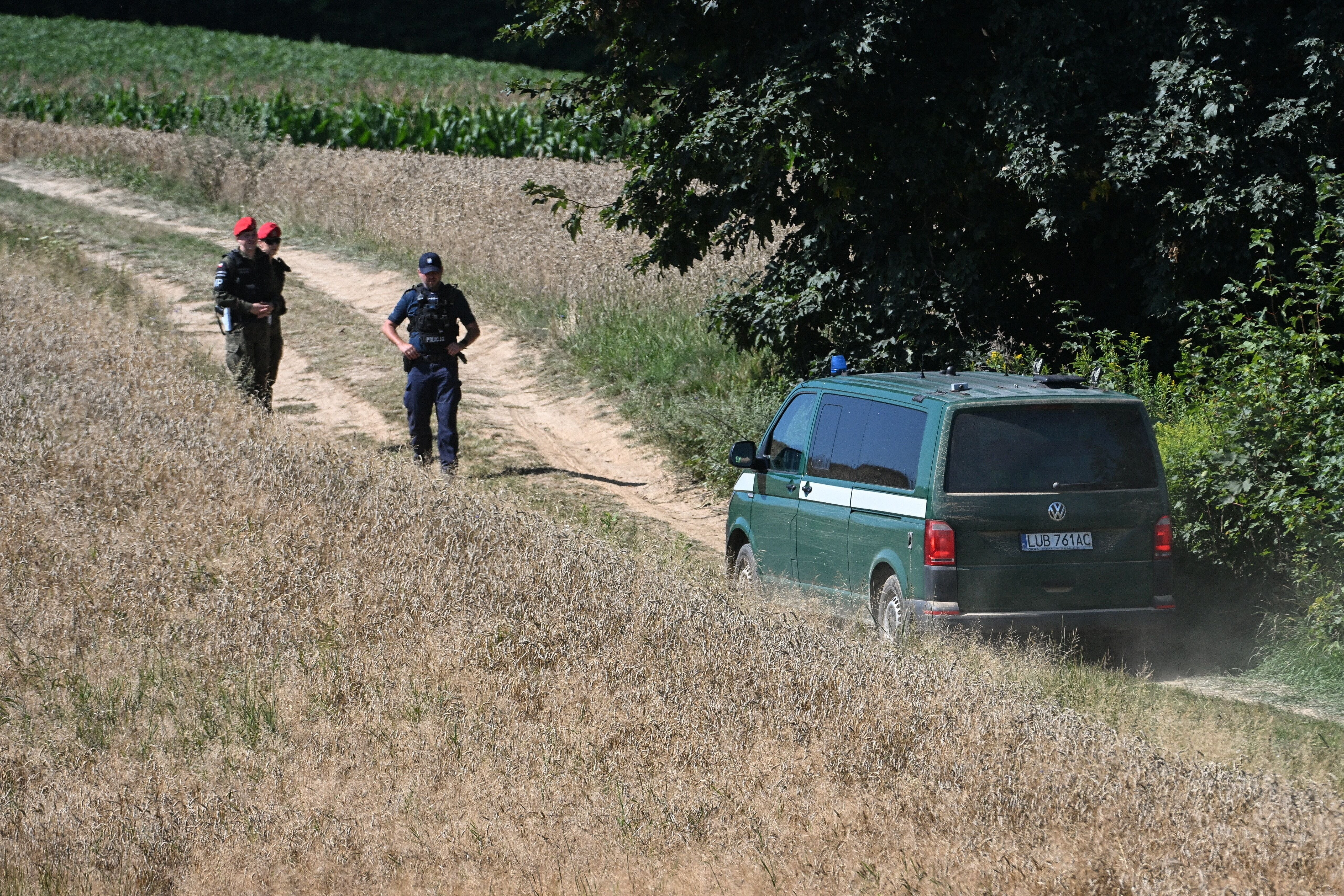 Polizisten und Soldaten gehen einen Feldweg entlang, neben ihnen steht ein grüner Einsatzwagen.