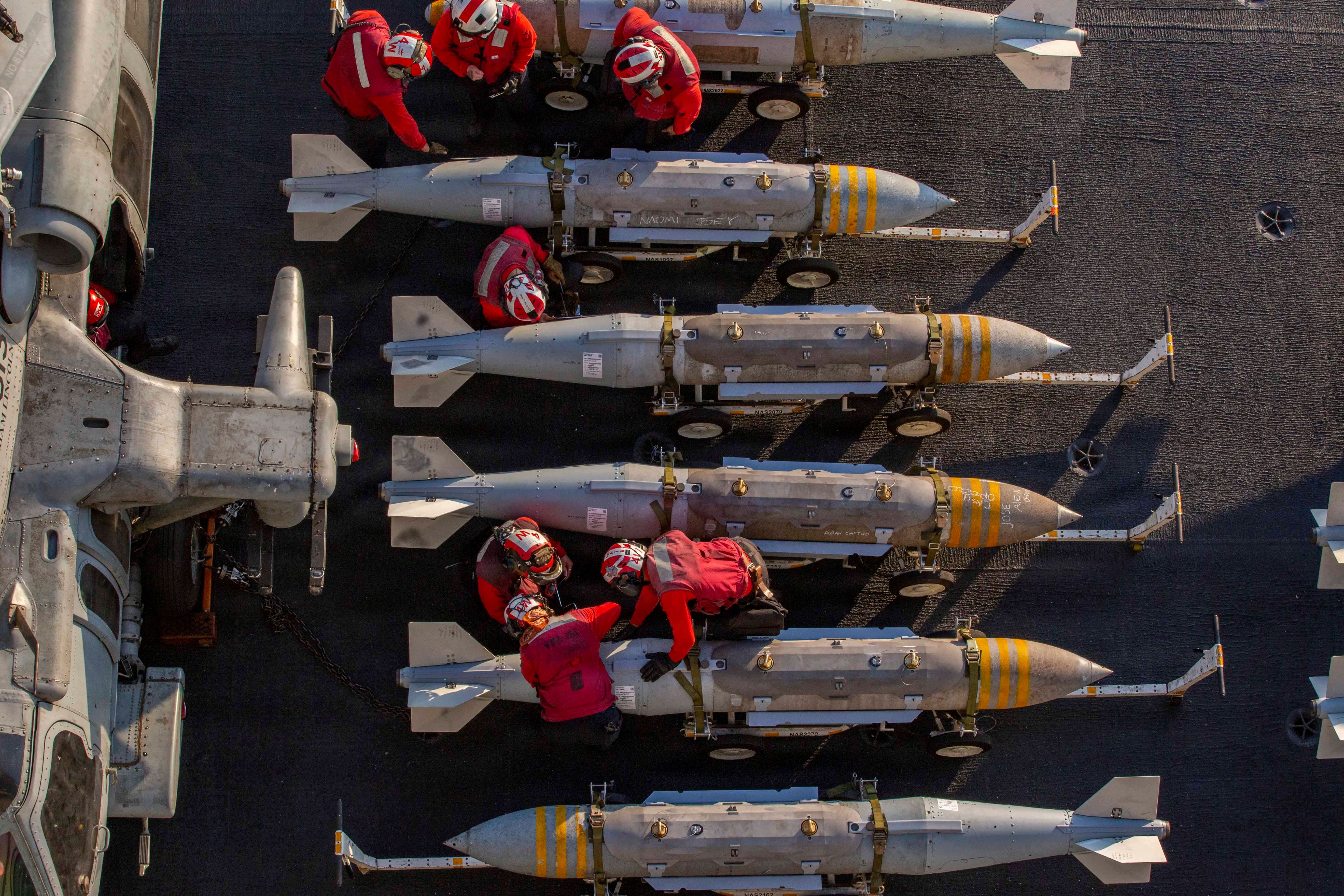 Mehrere US-Matrosen in roten Overalls bereiten Bomben auf Rollwagen auf dem Flugdeck eines Flugzeugträgers vor.