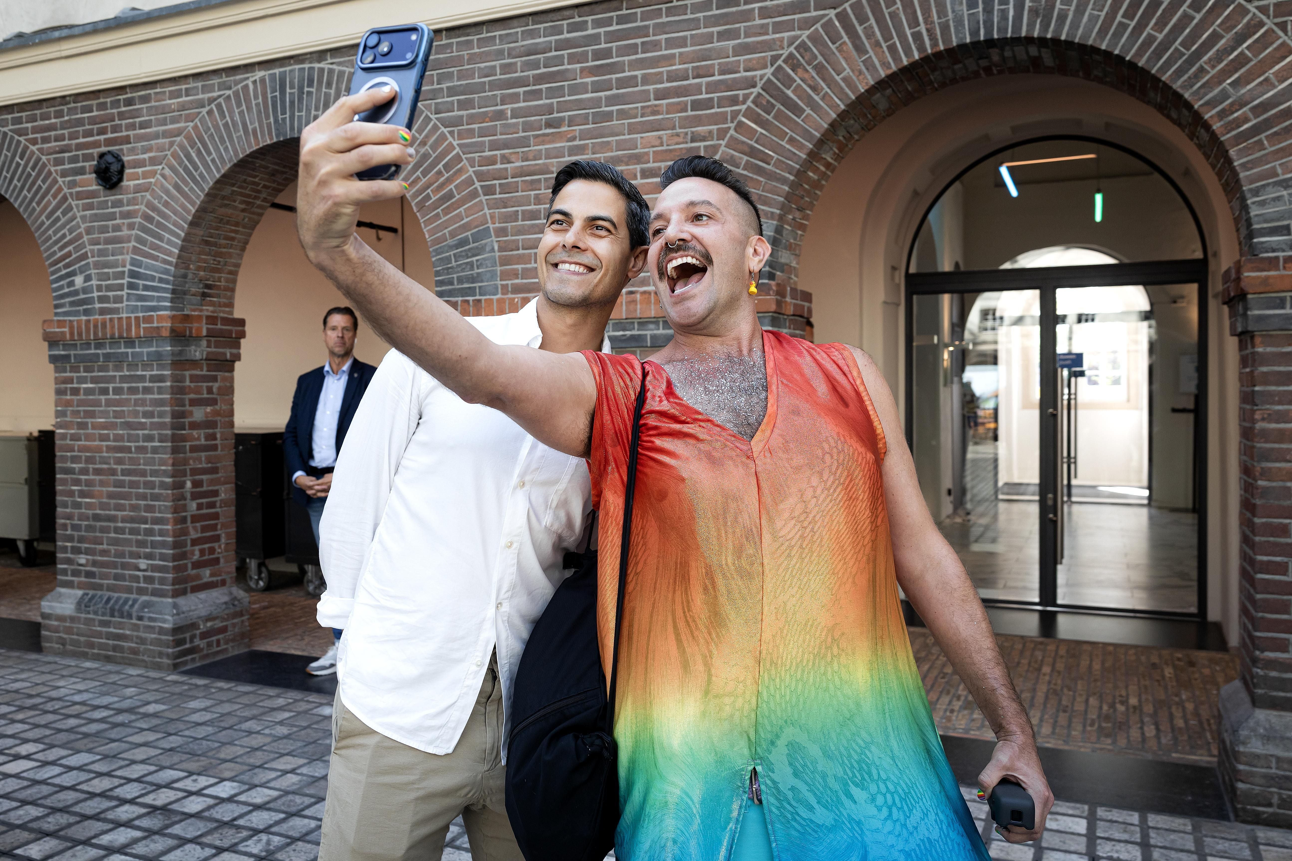 Zwei Männer machen ein Selfie bei Pride-Feierlichkeiten vor einem Backsteingebäude.