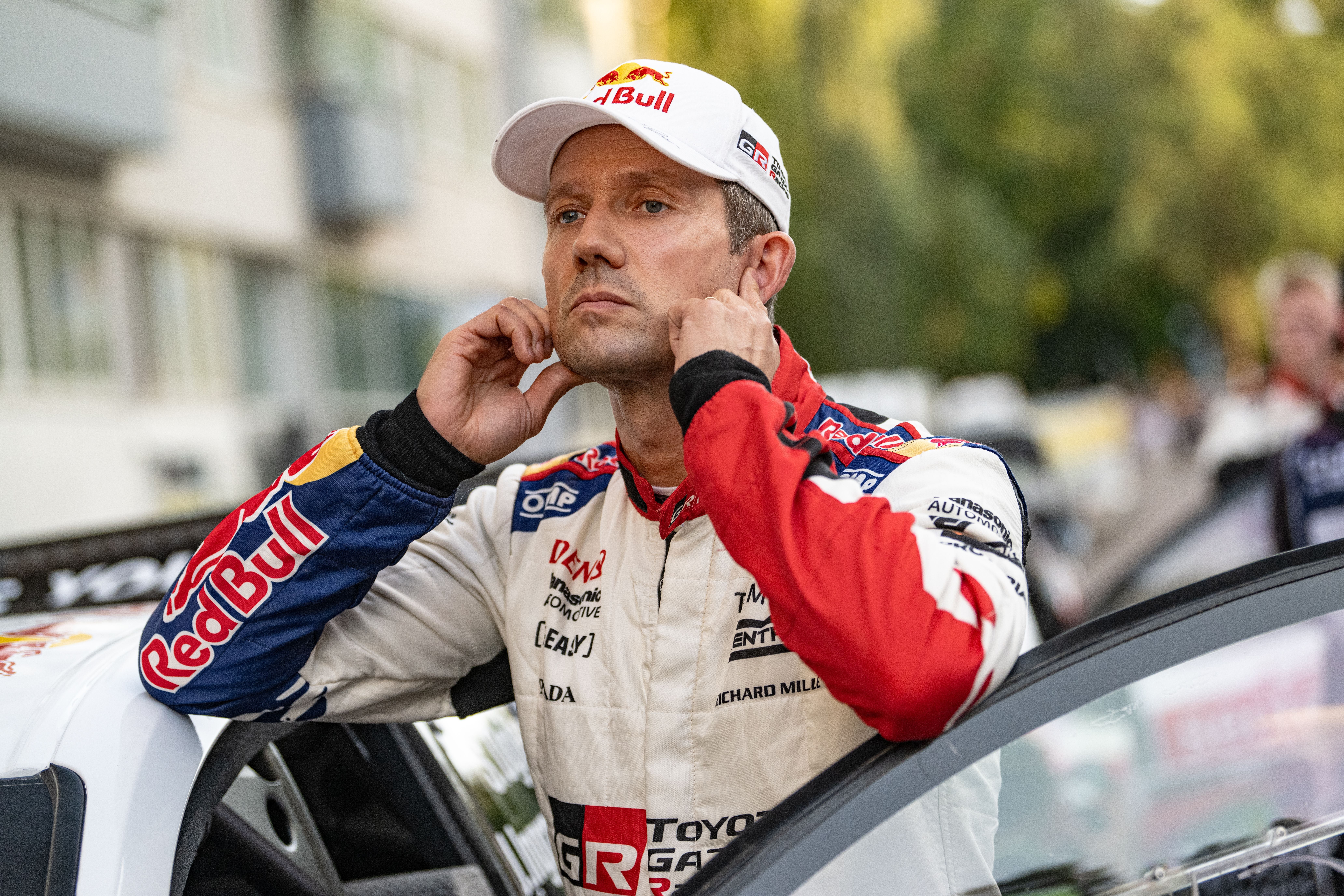 Ein Rallyefahrer im Rennanzug steht neben einem Auto und setzt sich Ohrstöpsel ein.