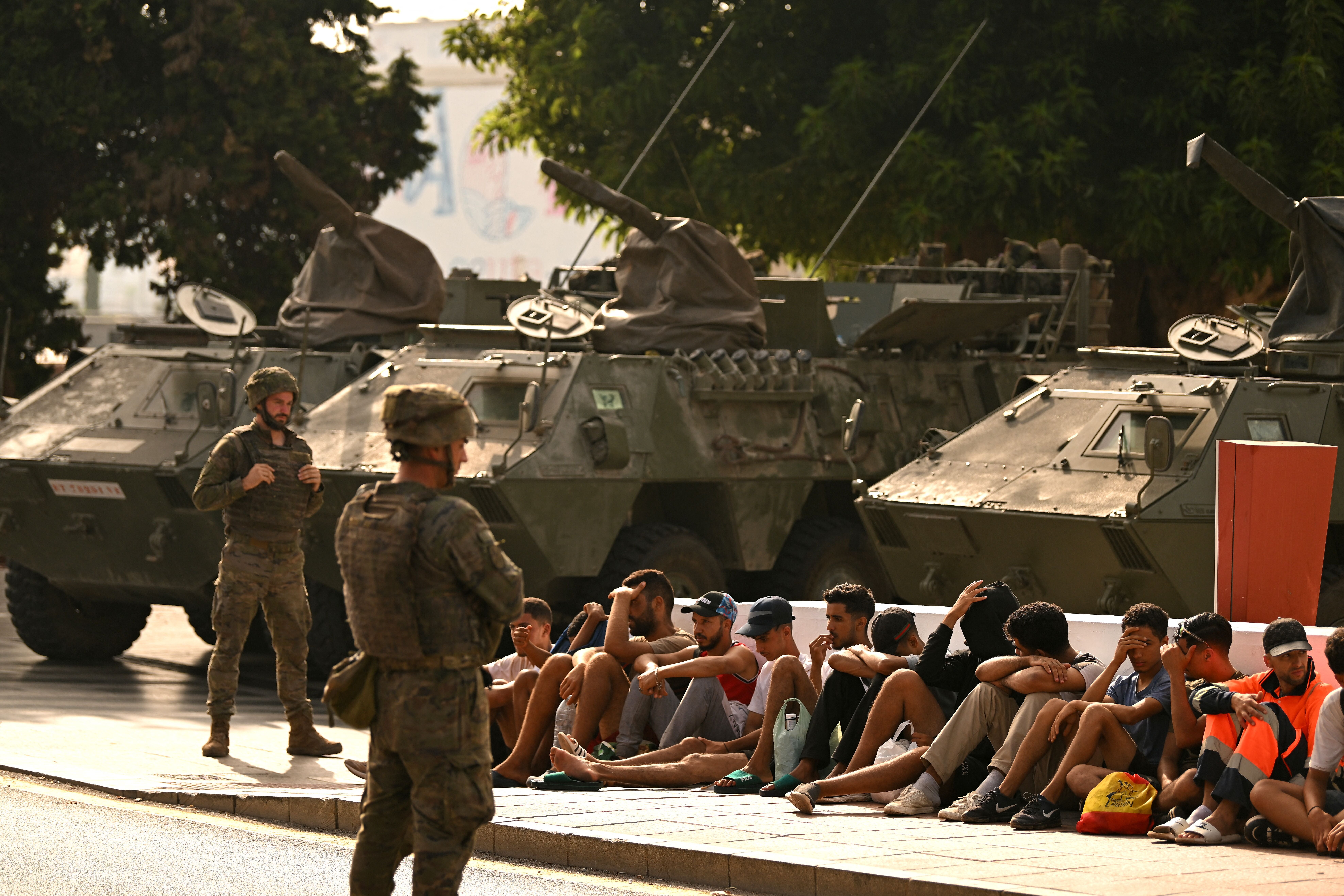 Spanische Soldaten bewachen sitzende Migranten vor Militärfahrzeugen in Ceuta.