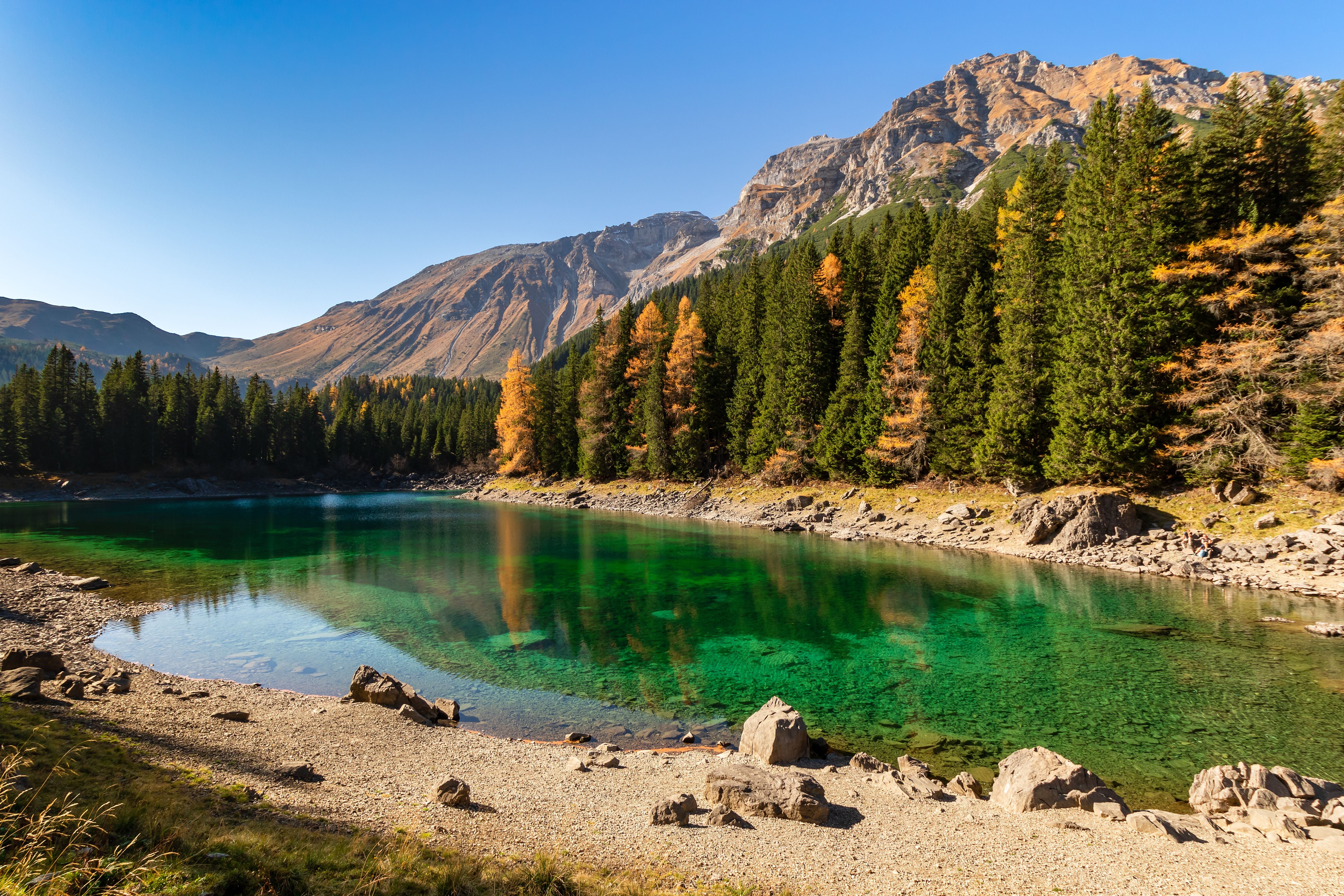 Bergsee mit grün-blauem Wasser, umgeben von Nadelbäumen und Bergen unter blauem Himmel.