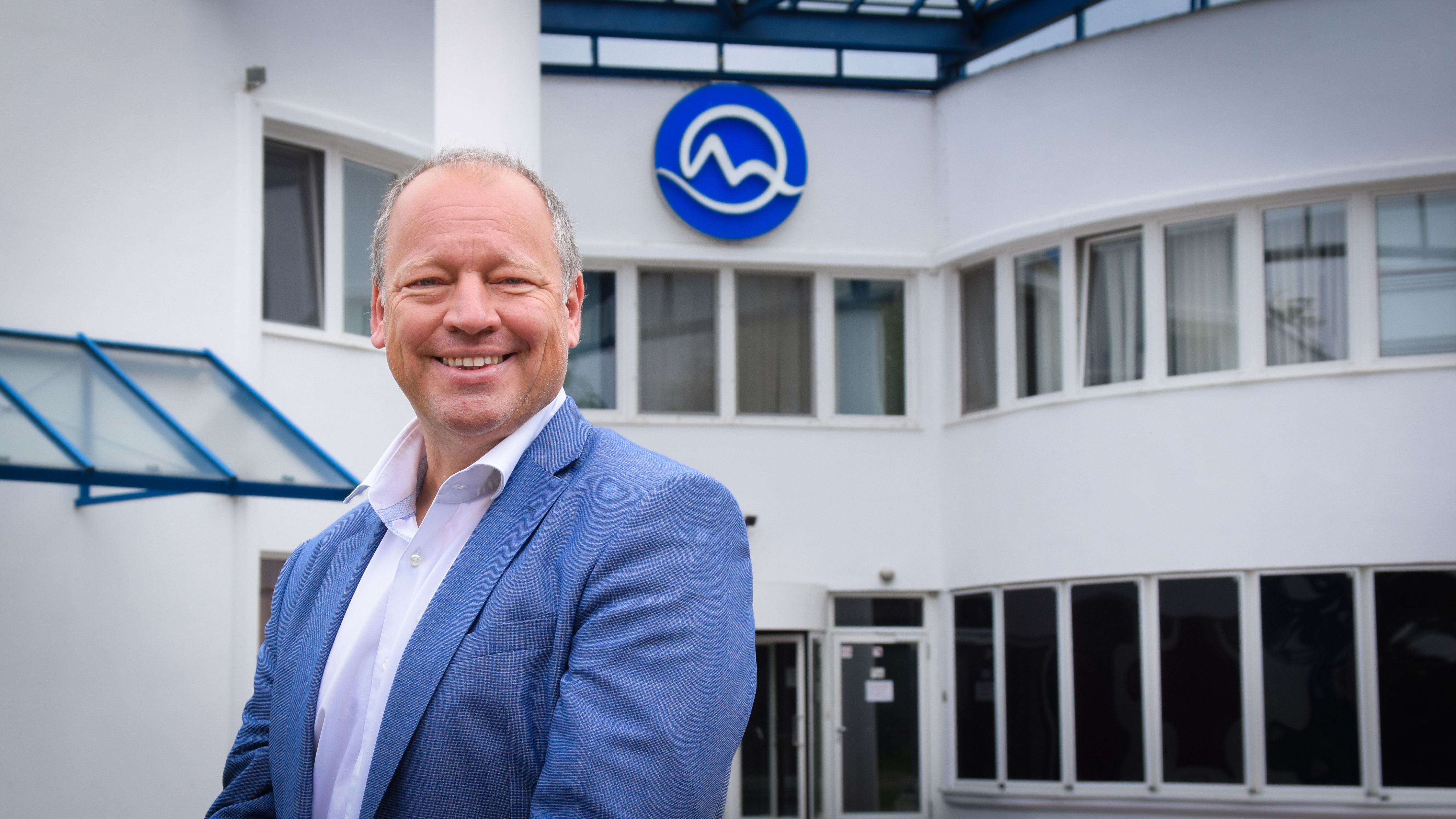 Ein Mann im blauen Sakko steht lächelnd vor einem modernen weißen Firmengebäude mit Logo.