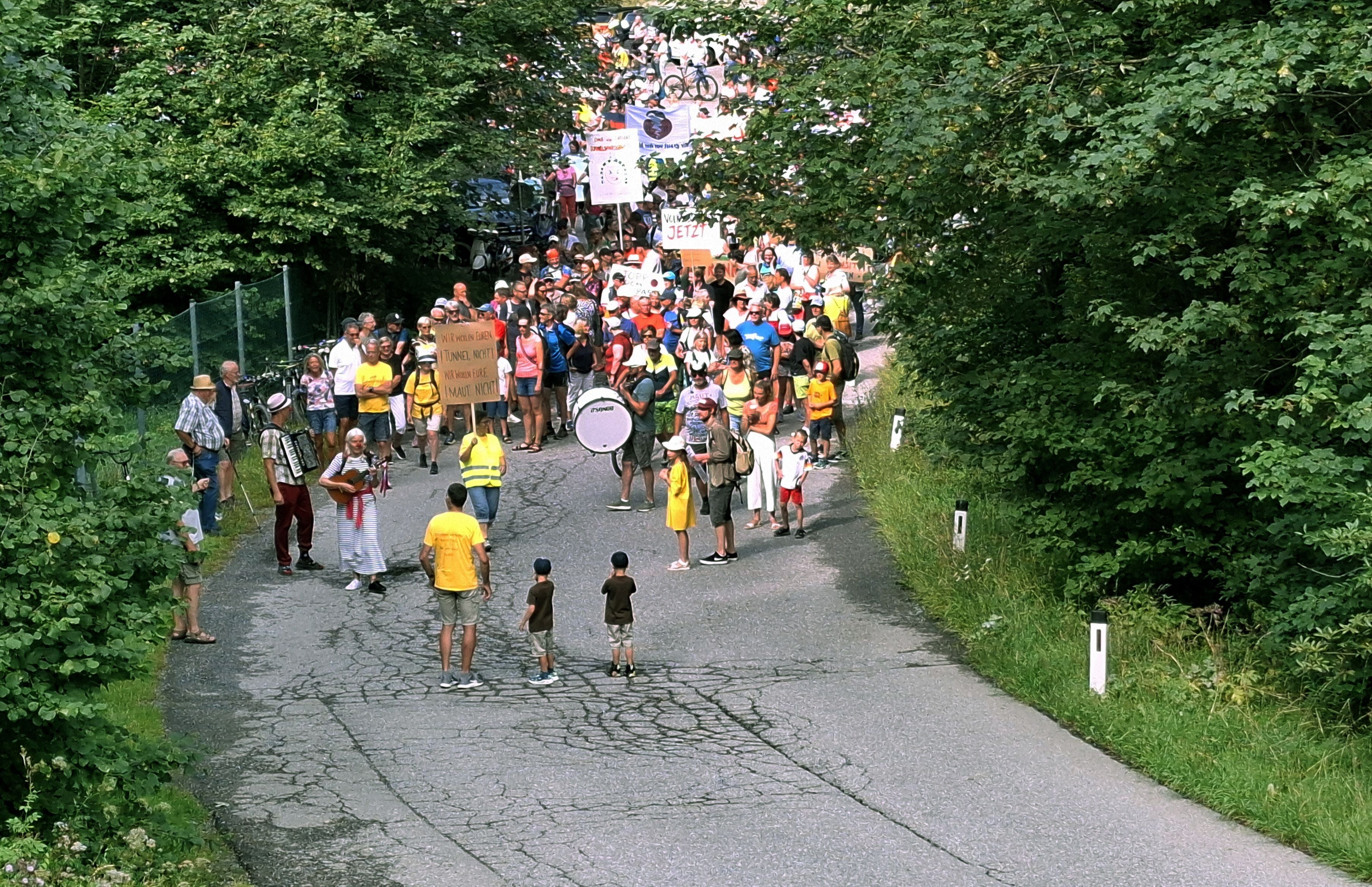 Eine große Gruppe von Demonstranten mit Schildern marschiert auf einer Landstraße durch bewaldetes Gebiet.