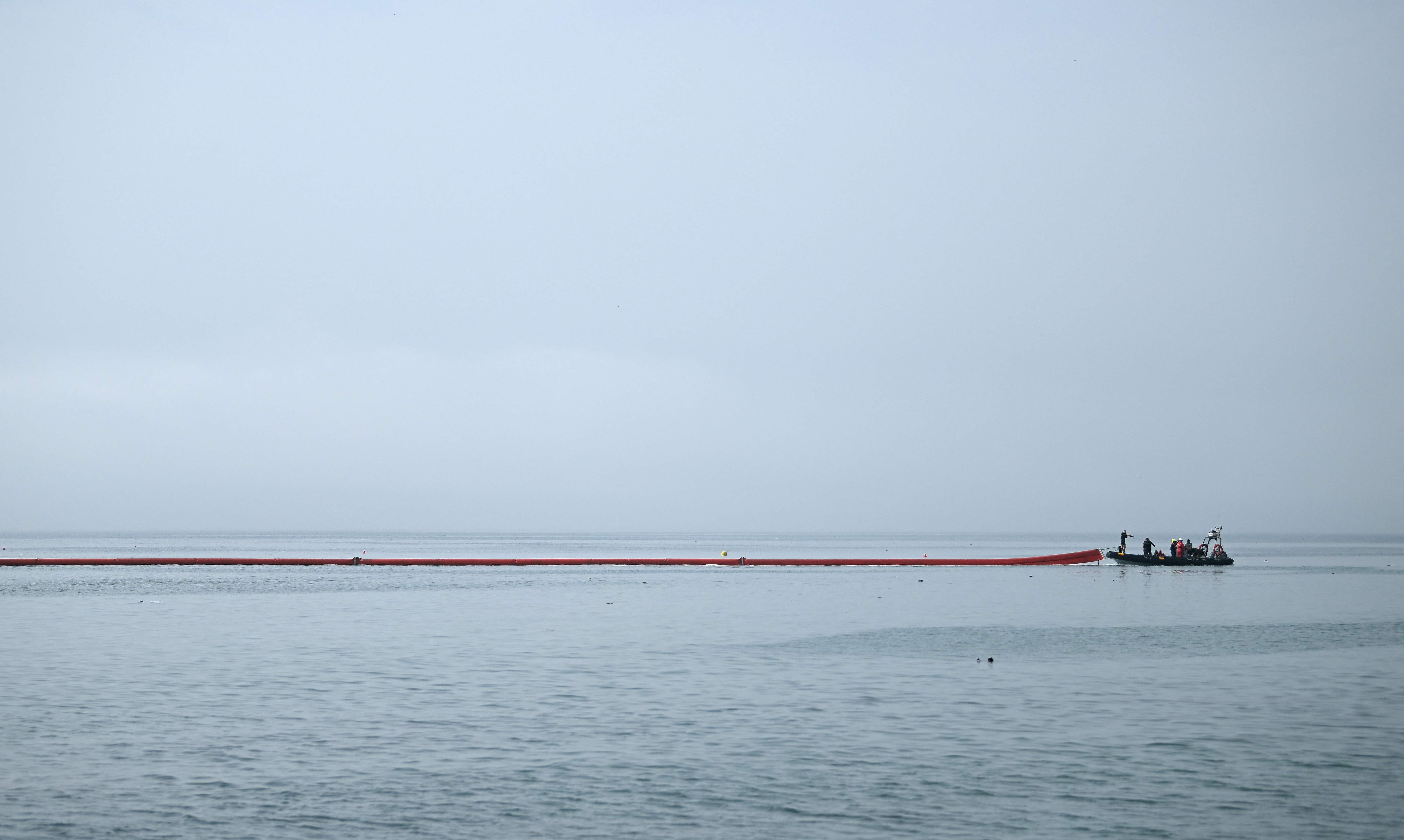 Ein langes rotes Barriereband wird von einem Boot auf ruhigem Meer ausgerollt.