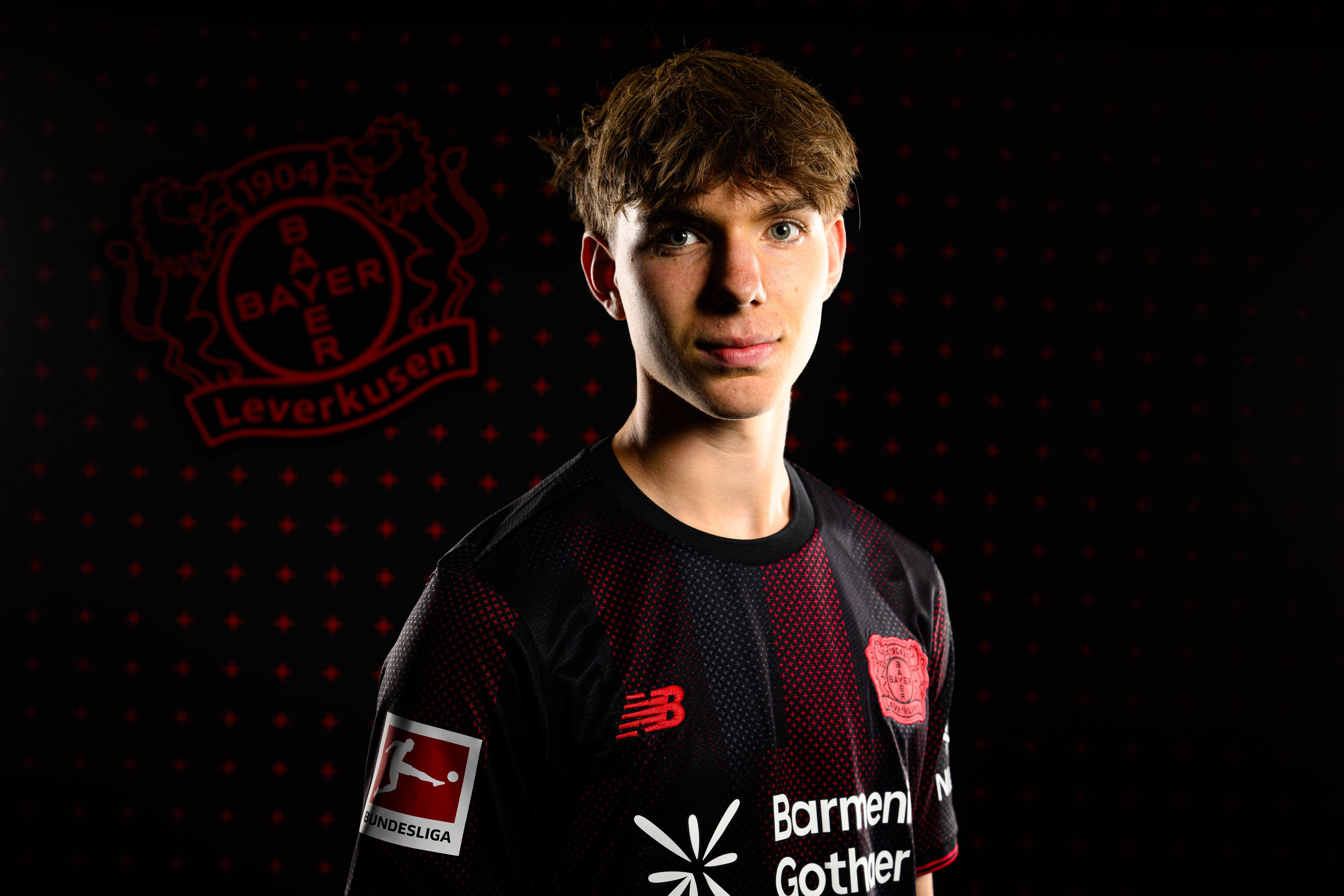 Ein junger Mann im Bayer-Leverkusen-Trikot posiert vor einem Vereinslogo auf schwarzem Hintergrund.